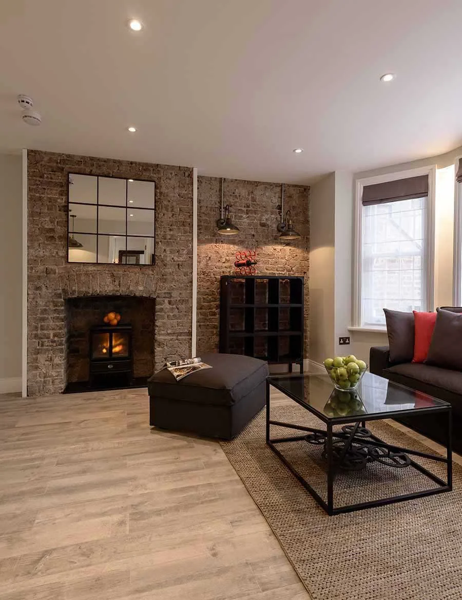 Interior photograph of a modern apartment living room at West End Lane, London, refurbished by Nick Baker Architects. The space features an original exposed brick wall integrating a fireplace.
