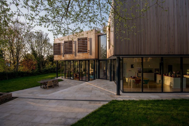Modern house with large glass windows, wooden exterior accents, and a paved patio with outdoor furniture, surrounded by trees and greenery.