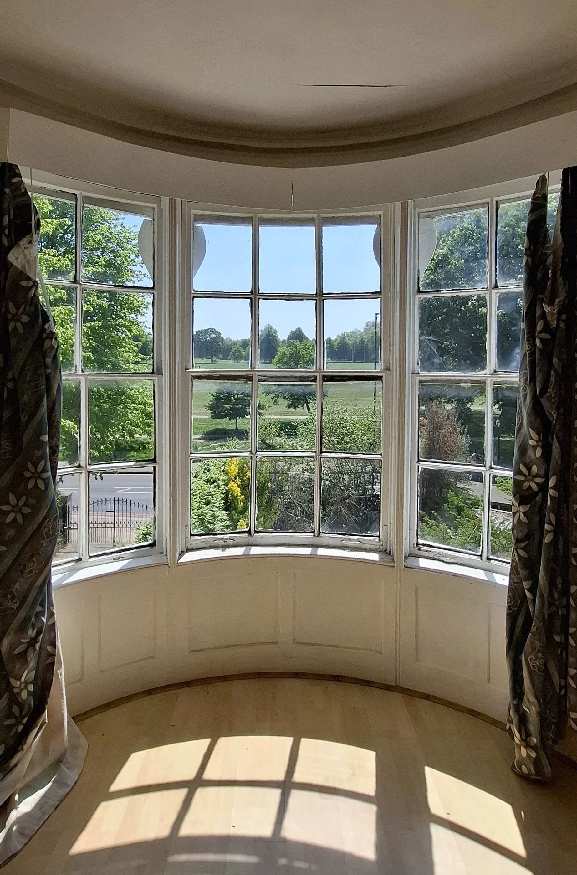 A large bay window with multiple panes, curtains drawn aside, overlooking a landscape with trees and a road.