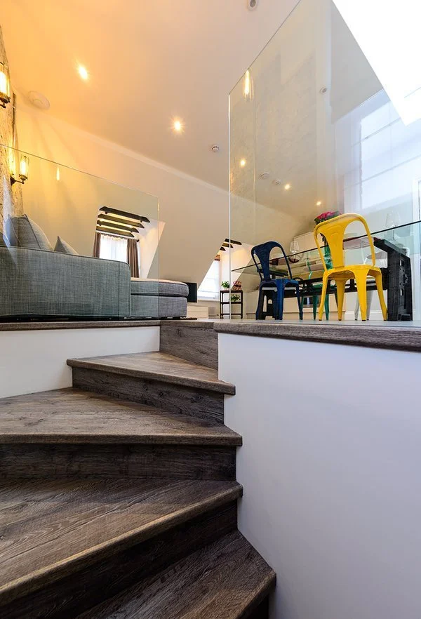 Interior of a modern living and dining area with dark wooden stairs, a gray sofa, a glass dining table, and colorful chairs, with natural light coming through large windows.