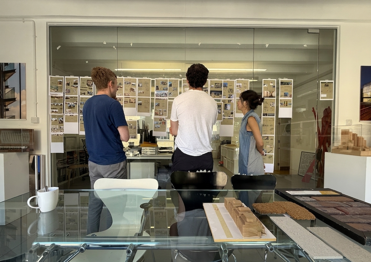 Three people are standing in front of display boards on a glass wall, looking at architectural or design plans. They are in a room with a glass table, a white mug, and various design samples and models.