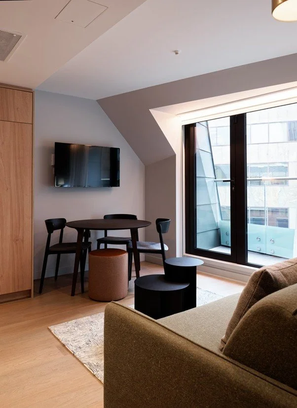 An interior view of an upper-floor hotel room suite at the Wilde Hotel. The space features a large black-framed glass doors leading onto a balcony framed by the building's angled mansard roofline.