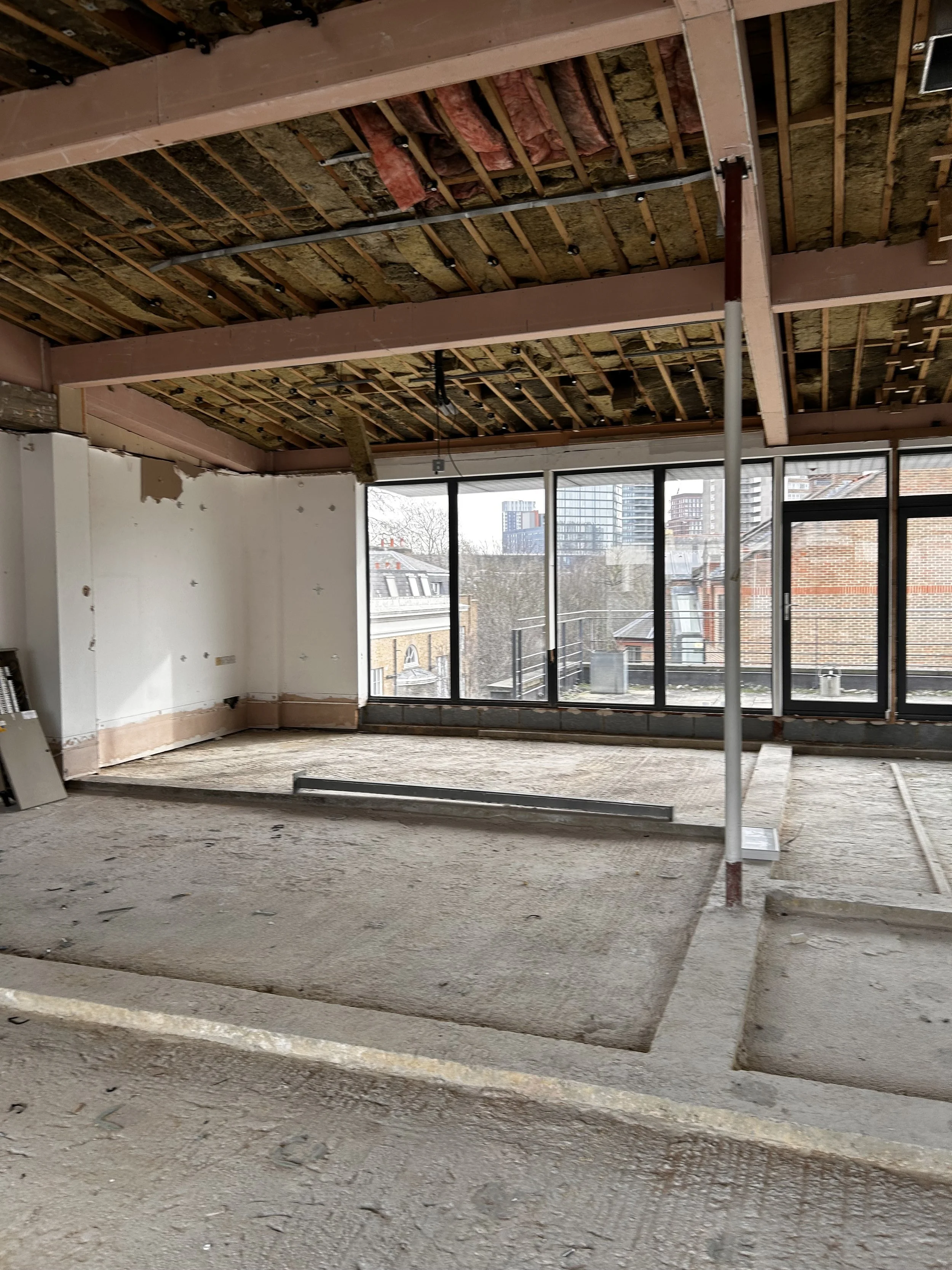 Interior of a building under construction with exposed ceiling framing, insulation, large windows, and a concrete floor.