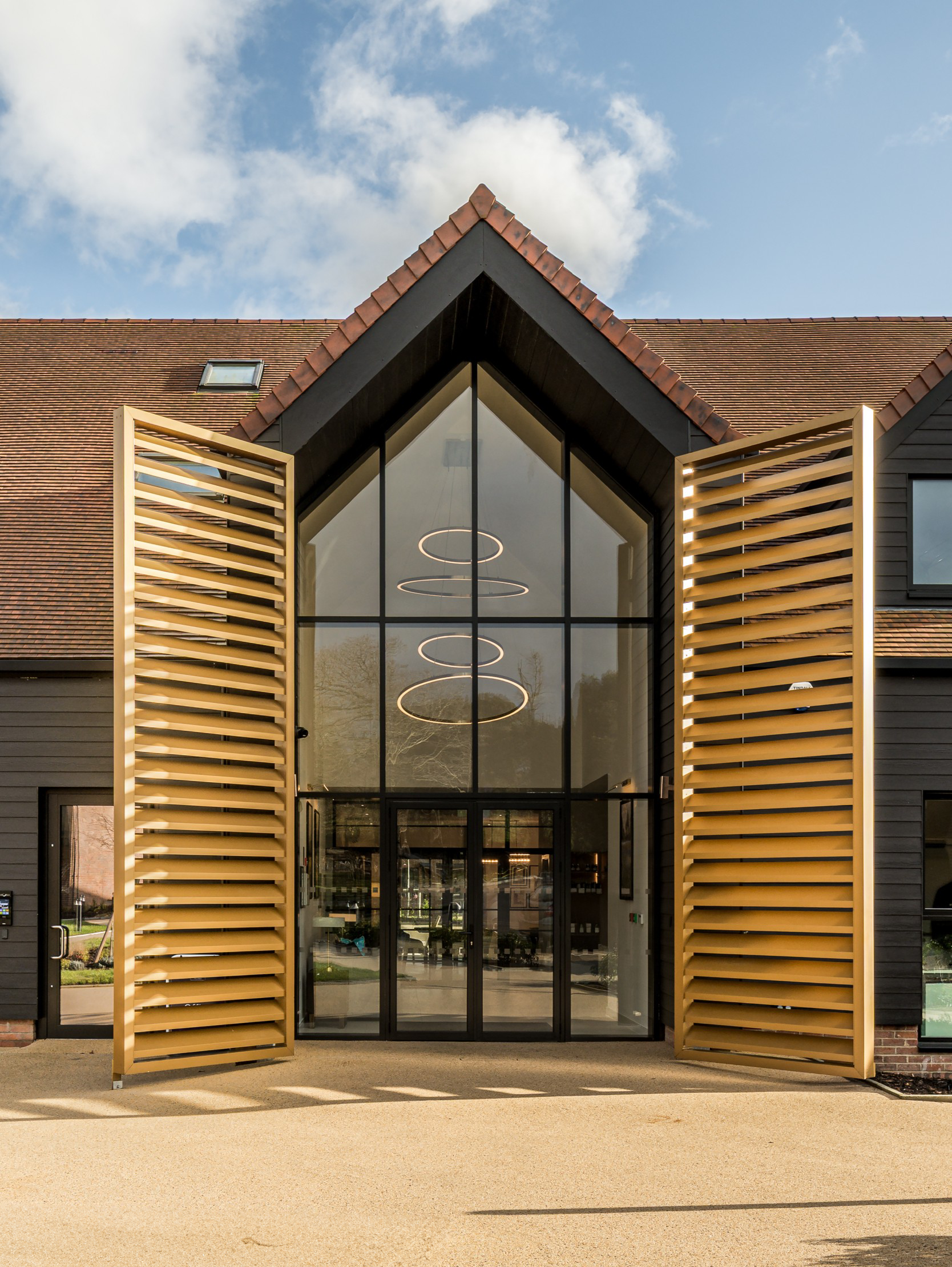 Modern barn-style clubhouse main entrance featuring a grand double-height glazed gable facade, dark timber cladding, and prominent gold external louvred door on a sunny day.
