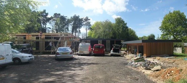 Construction site with partially built wooden structures, parked cars, and construction materials on the ground under a blue sky with clouds.