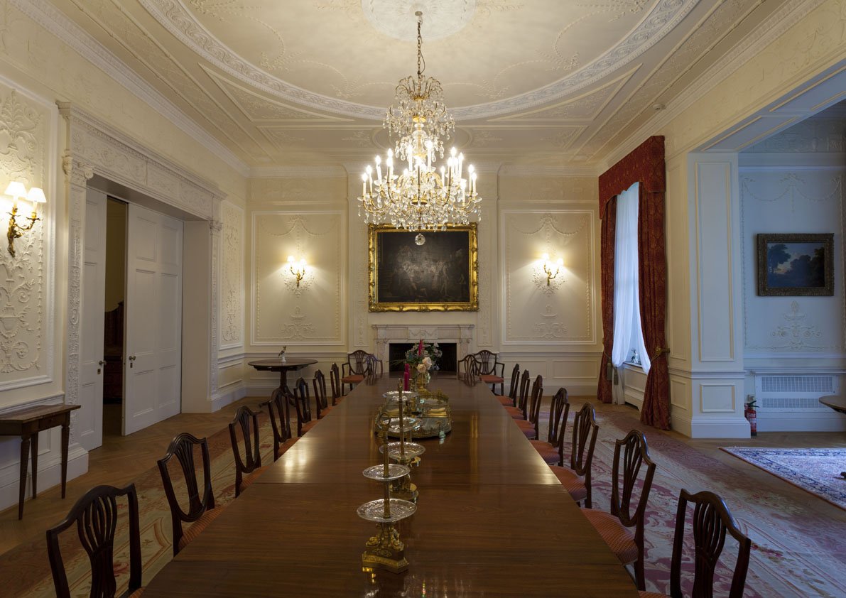 Elegant oval dining room with a long wooden table, dozens of chairs, ornate white walls, a large chandelier hanging from the ceiling, gold-framed portrait above the fireplace, and windows with red curtains.