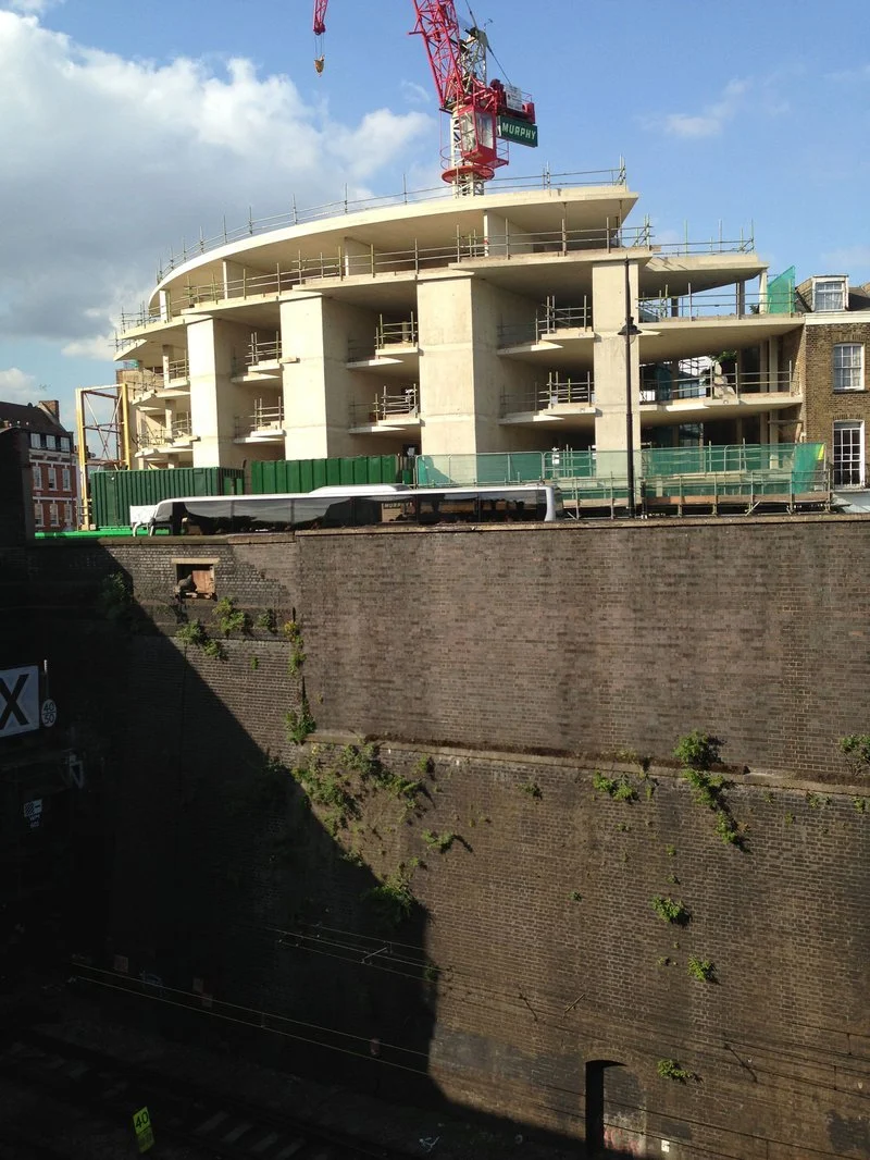 Construction site with a multi-story building under construction, crane overhead, and a train passing by on a railway below a brick wall.