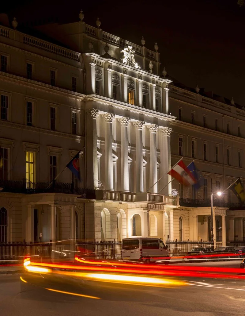 Night view of a grand, illuminated building with neoclassical architecture, featuring tall columns and a crest on top, with flags hanging from the facade and light trails from passing vehicles in front.