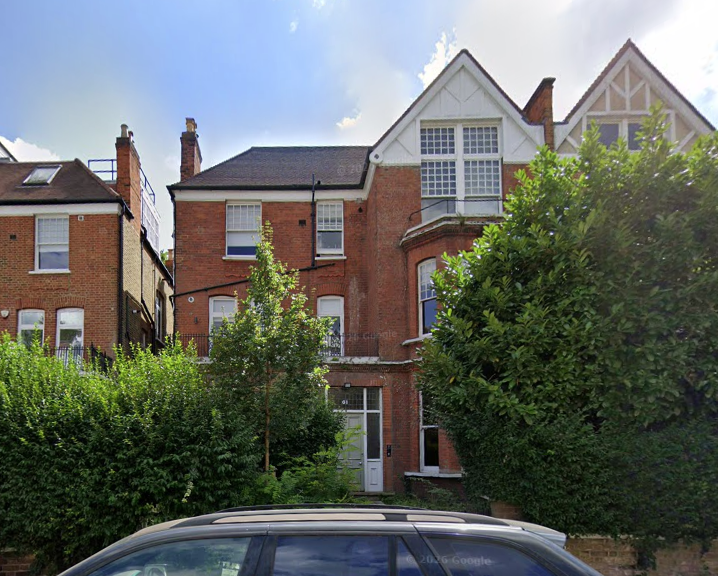 A multi-story brick house with white trim and large windows, surrounded by green trees and bushes, with a car parked in front.