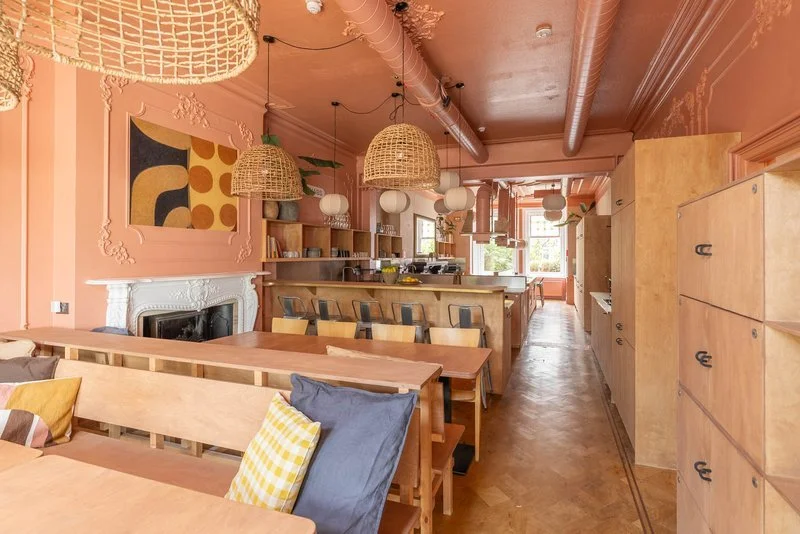 A cozy cafe interior with pink walls, wooden furniture, pendant wicker and globe lights, a fireplace, and colorful cushions on the seating area.