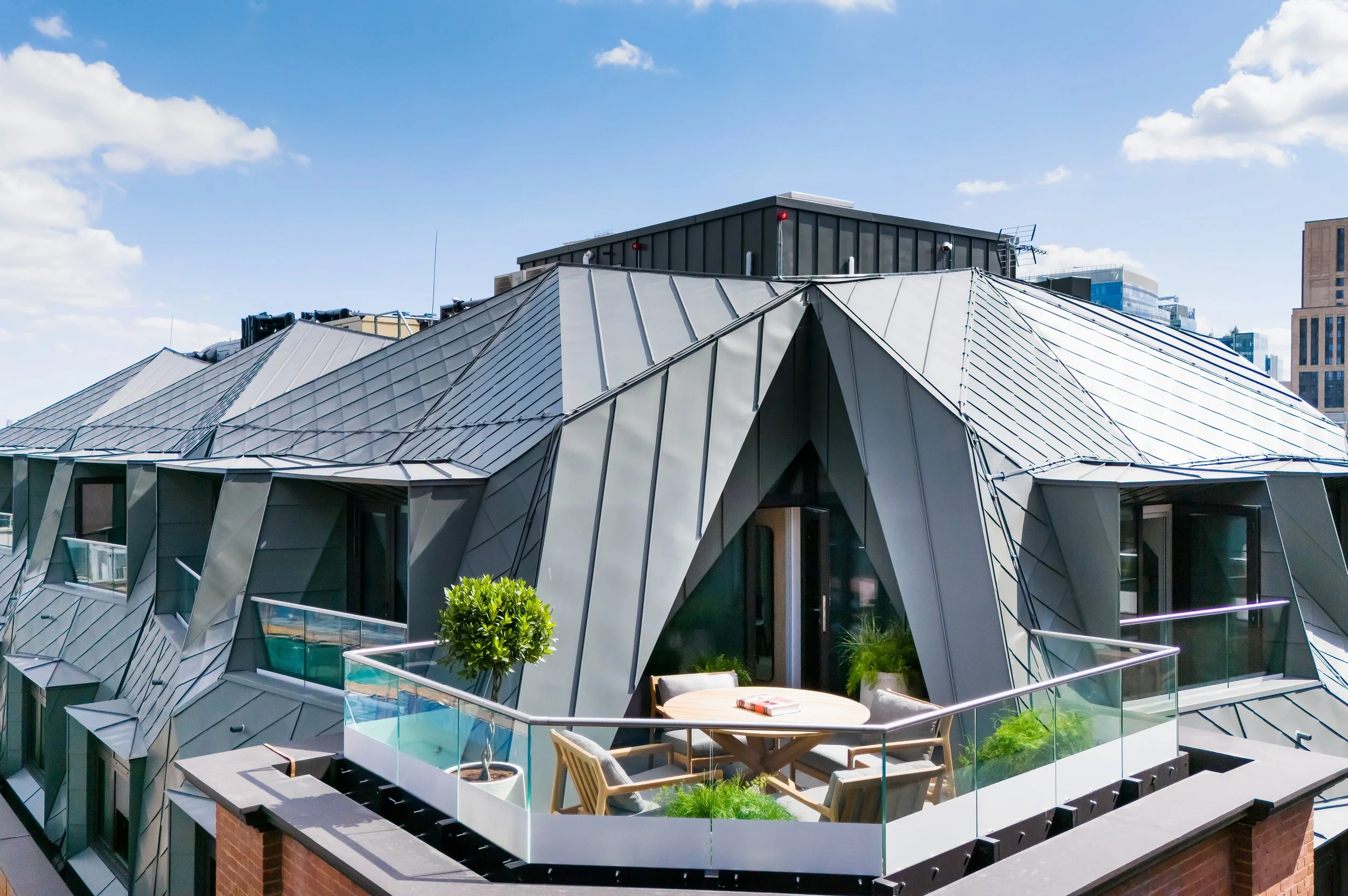 A view looking along a guest balcony at the Wilde Hotel roof extension. The terrace features a gray standing-seam zinc-clad mansard structure with deep, angular geometric facets framing large glass doors with a cityscape in the background.