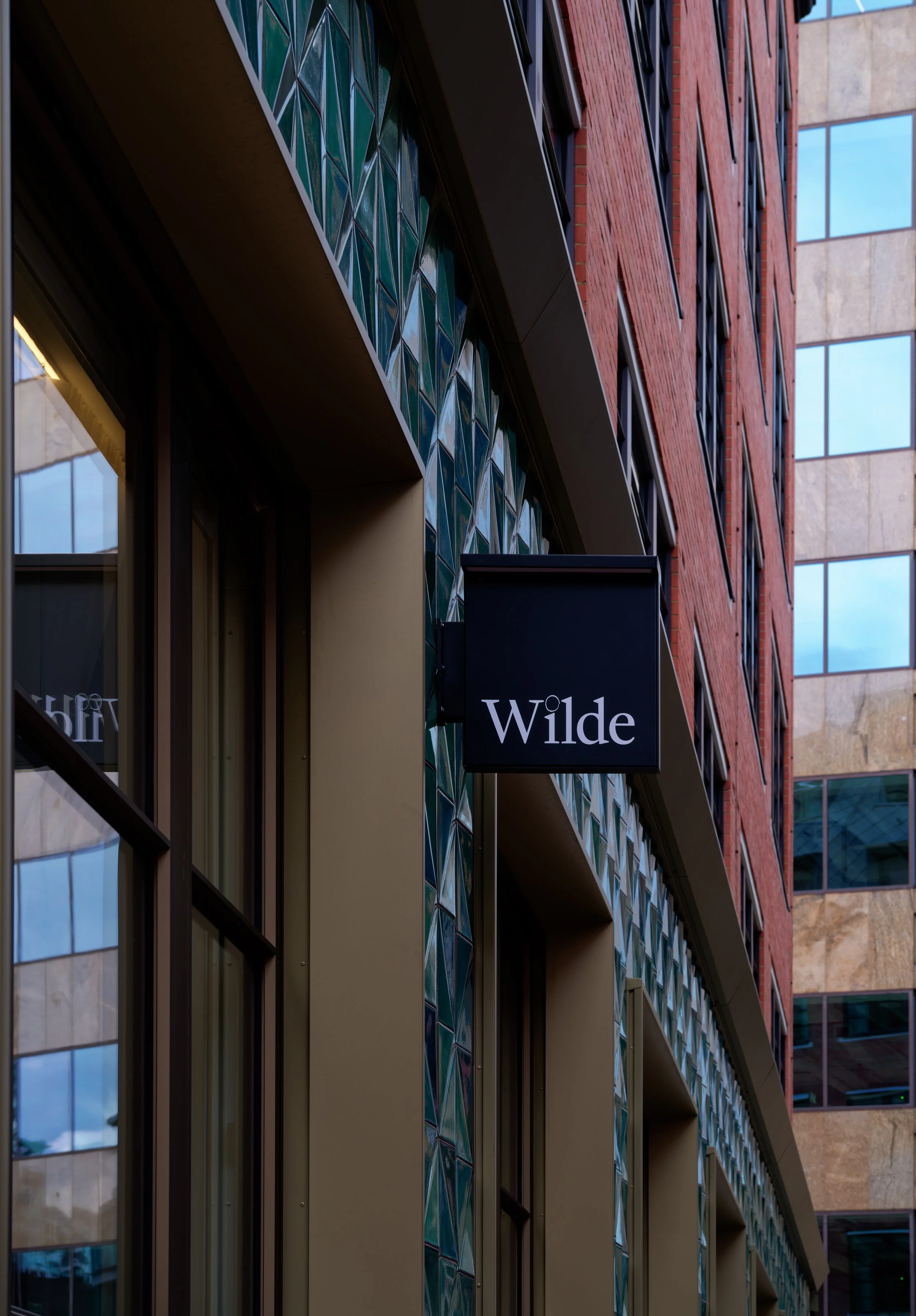 A low-angle street view looking along the facade of the Wilde Hotel on Strype Street. The shot highlights the sharp, contemporary bronze window profiles