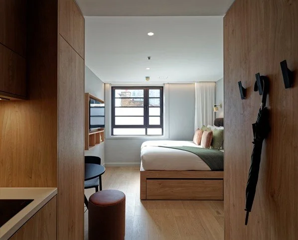 A bright, modern hotel room interior at the Wilde Hotel viewed from an entryway flanked by warm oak-paneled cabinetry.