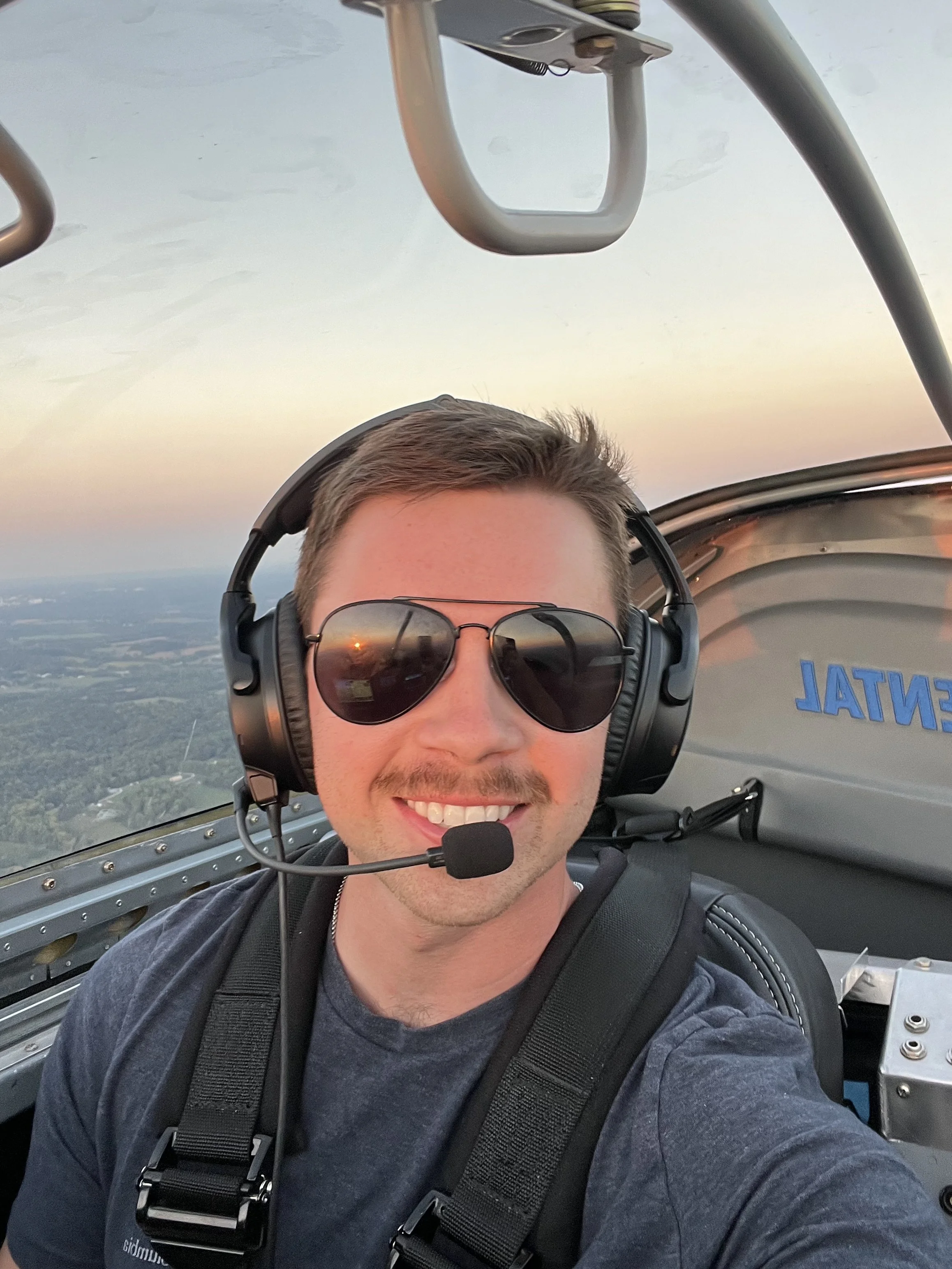 A young man flying a small aircraft, wearing sunglasses, a headset with microphone, and a black backpack, smiling at the camera with a sunset sky and landscape in the background.