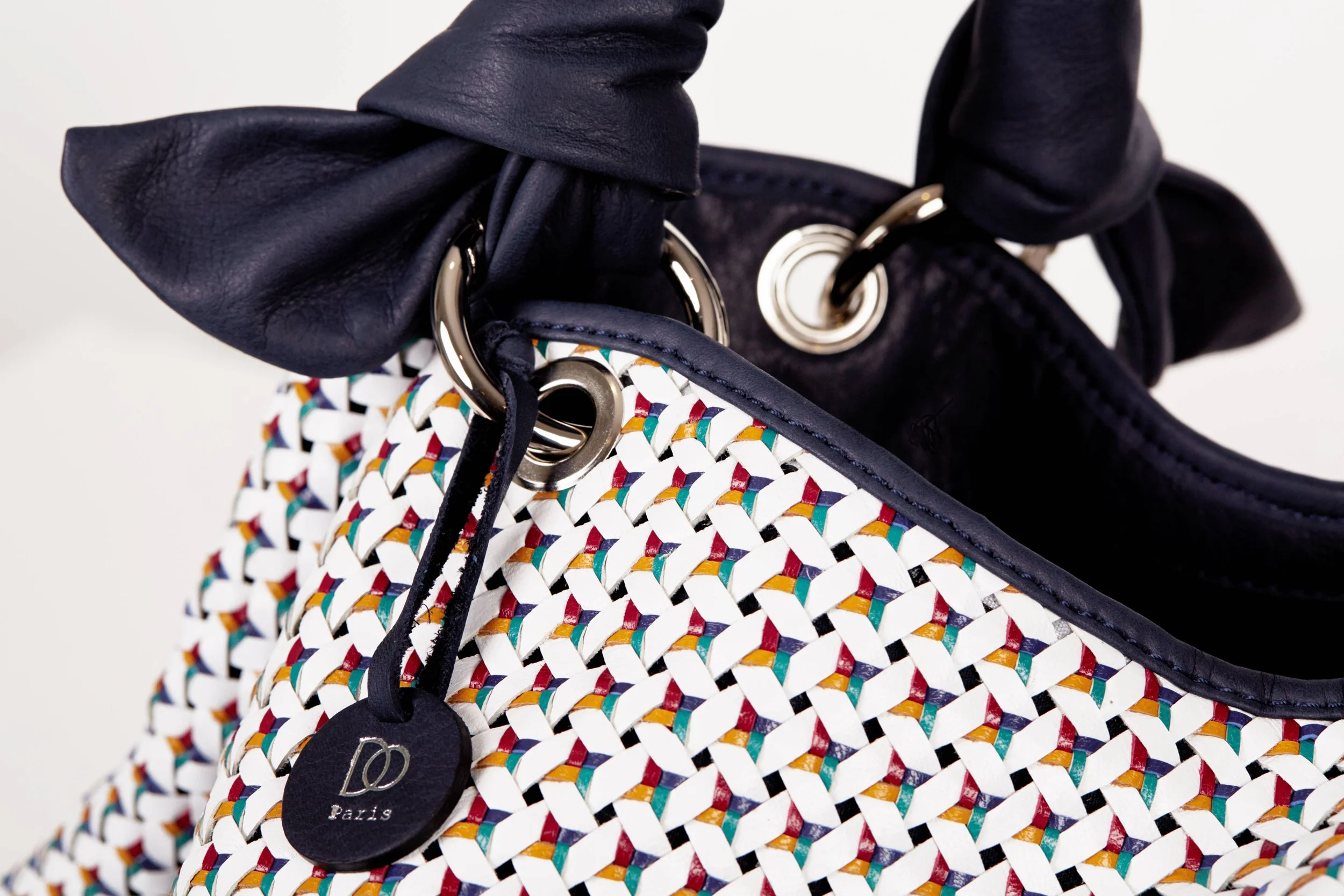 Sac à main en cuir noir avec une poignée en métal, attaché à une chaussure en tissu coloré à motif en entrelacs, avec une étiquette Paris.