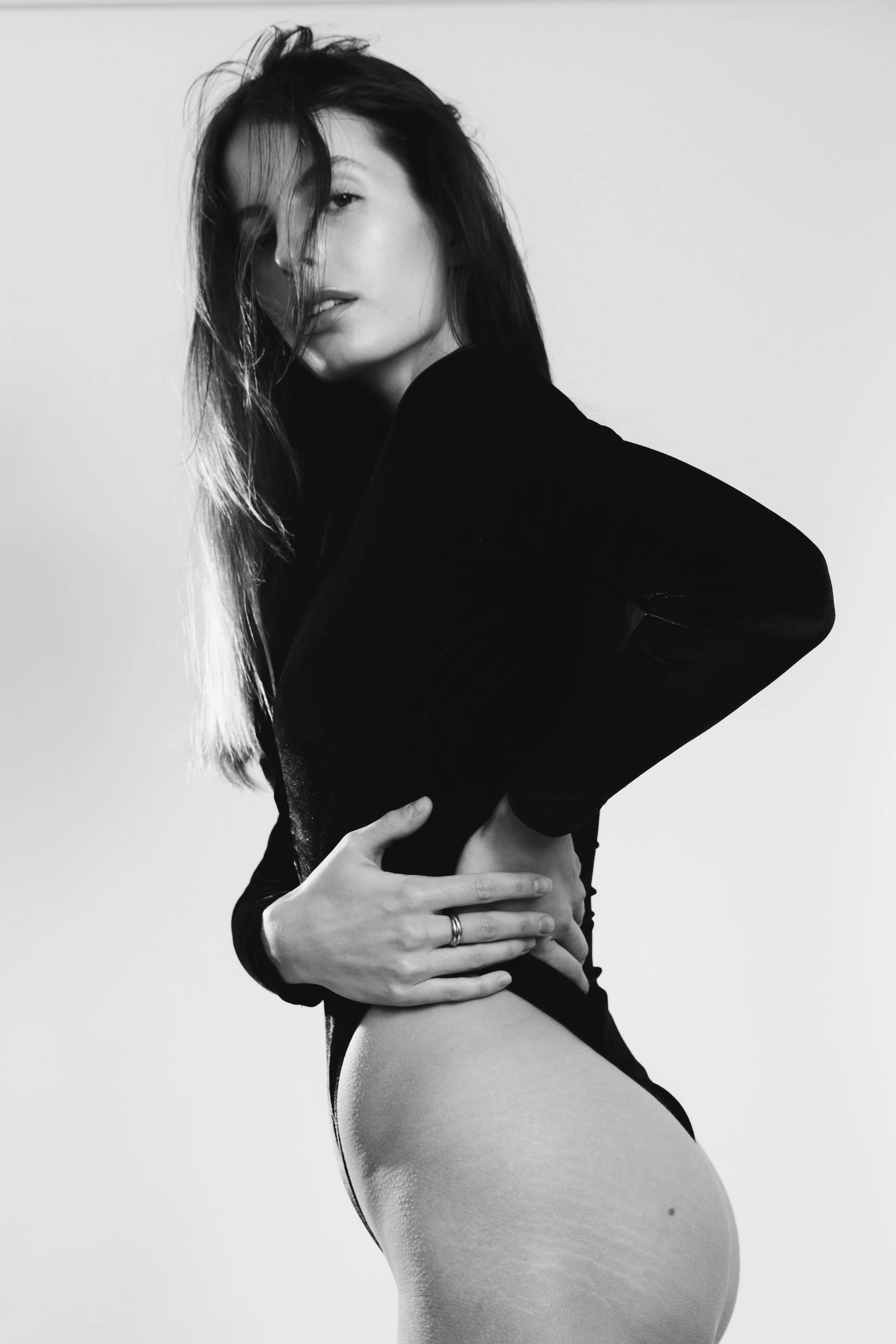 Une femme en noir et blanc pose avec ses cheveux en désordre, portant un vêtement noir, avec une main posée sur sa taille et l'autre sur sa cuisse, en regardant vers l'objectif, dans un style artistique et sophistiqué.