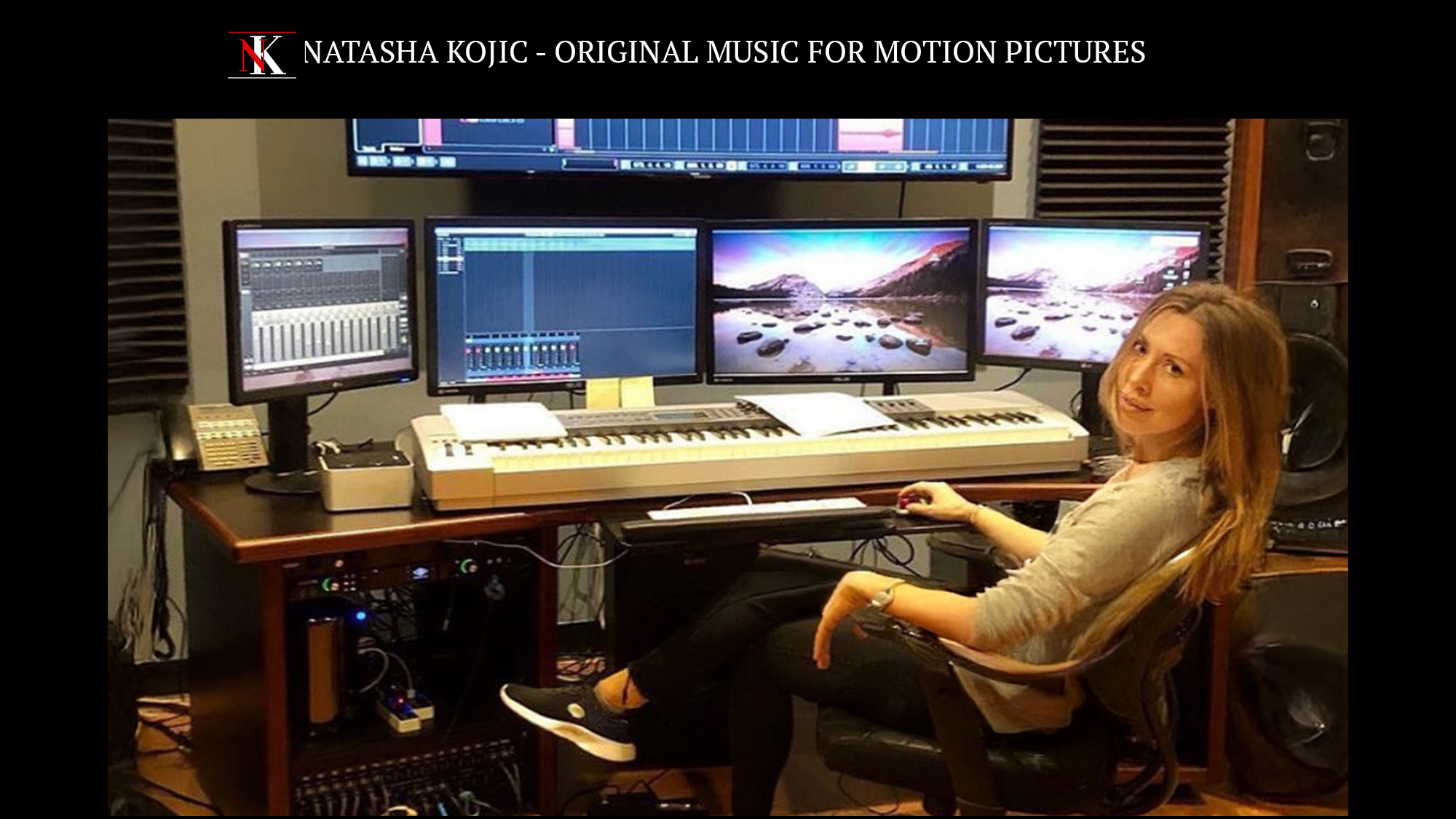 A woman sitting in a music studio with multiple monitors, a keyboard, and audio equipment, working on digital music production.