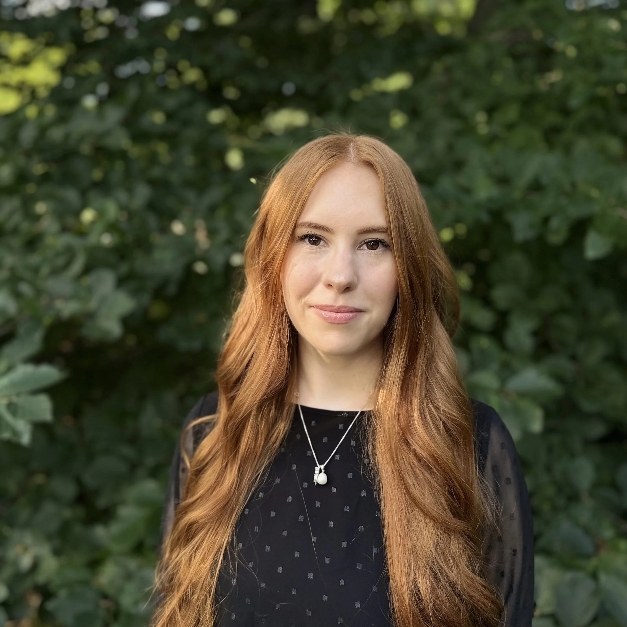 Close-up of a woman with long, wavy red hair, smiling softly, standing outdoors in front of green foliage.