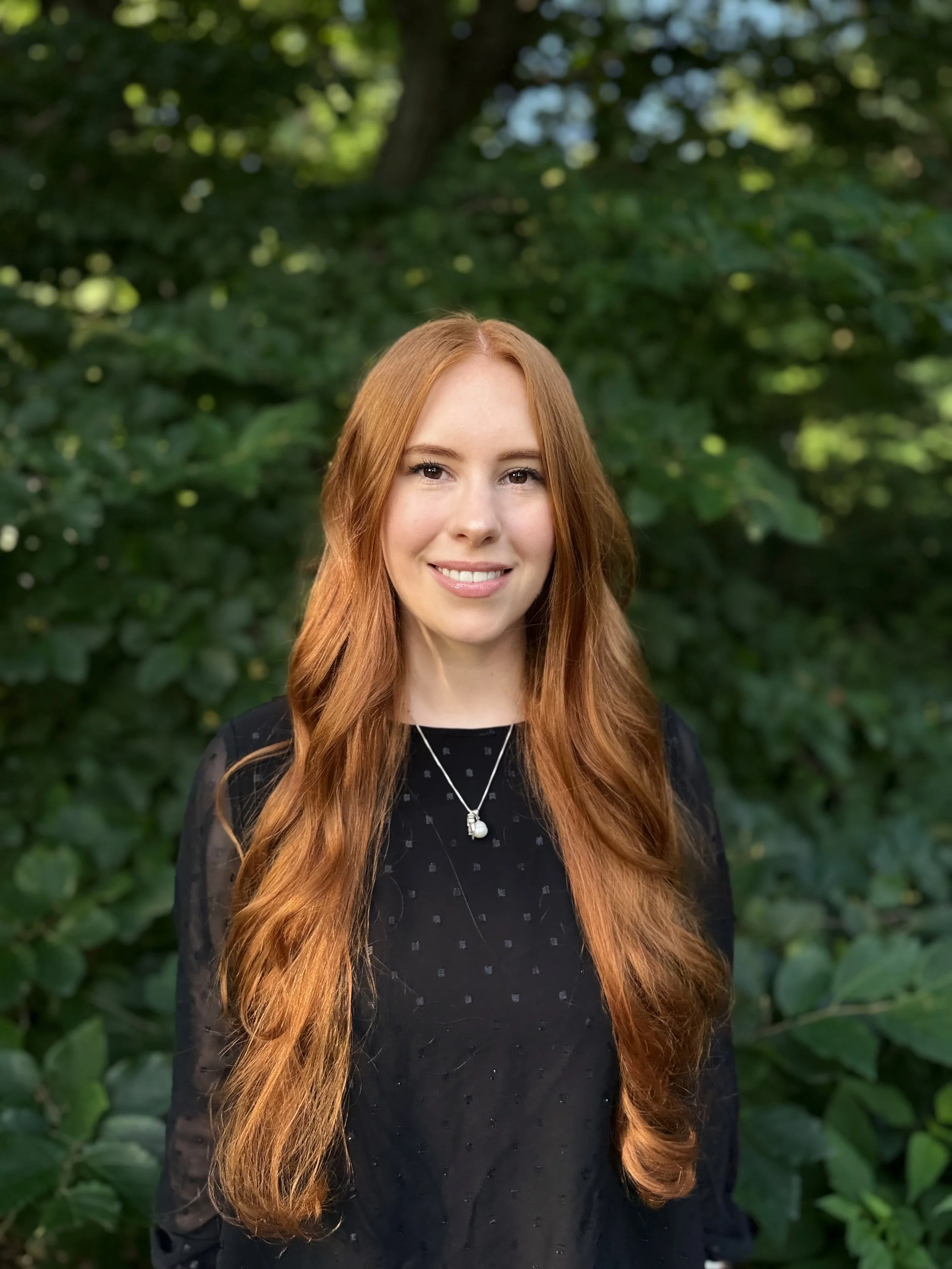 Portrait of a woman with long, wavy red hair, smiling, outdoors with green foliage in the background.