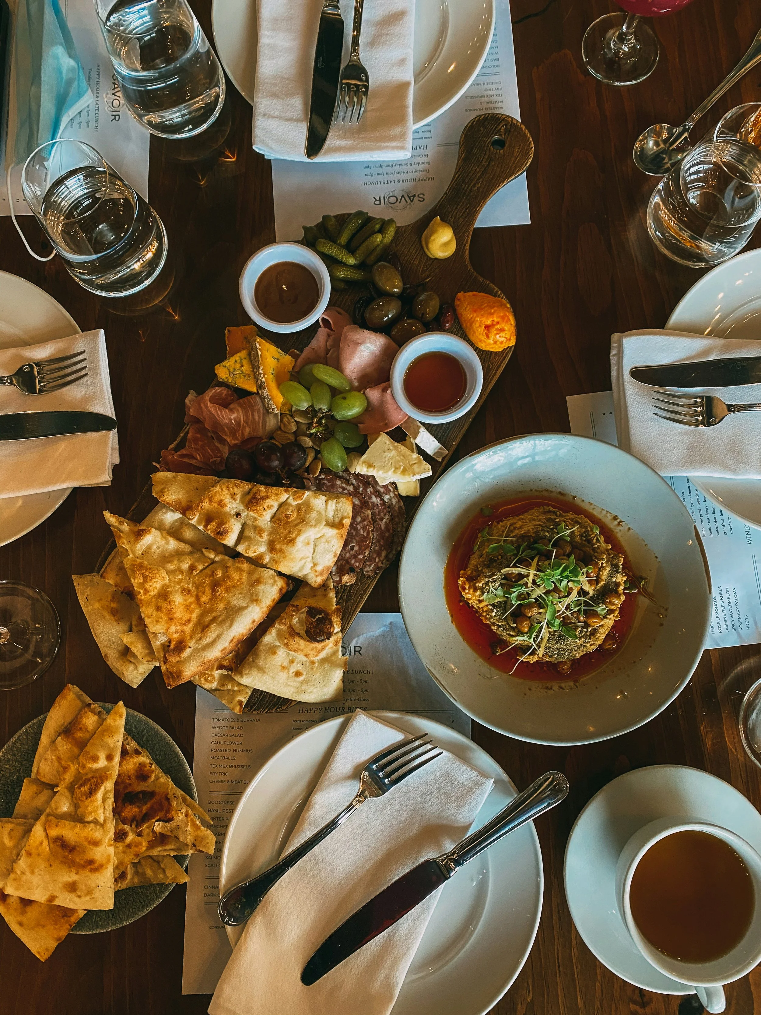 A table set for a meal with various dishes including a charcuterie board with meats, cheeses, grapes, and pickles, slices of pizza, a bowl of stuffed eggplant, and multiple glasses of water, wine, and coffee. There are plates, napkins, utensils, and menus around the table.