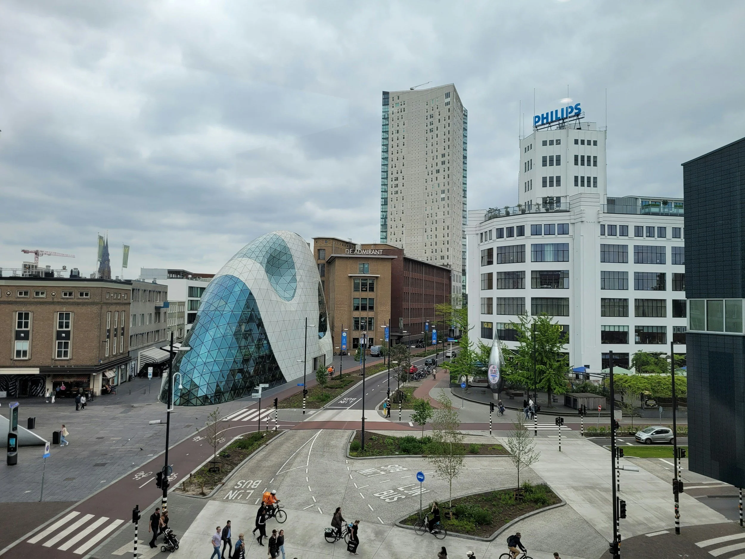 Stadsplein met moderne gebouwen, inclusief een uniek architectonisch gebouw met een boogvorm en glasoppervlak, mensen die lopen en fietsen, en aangeplante groene gebieden.