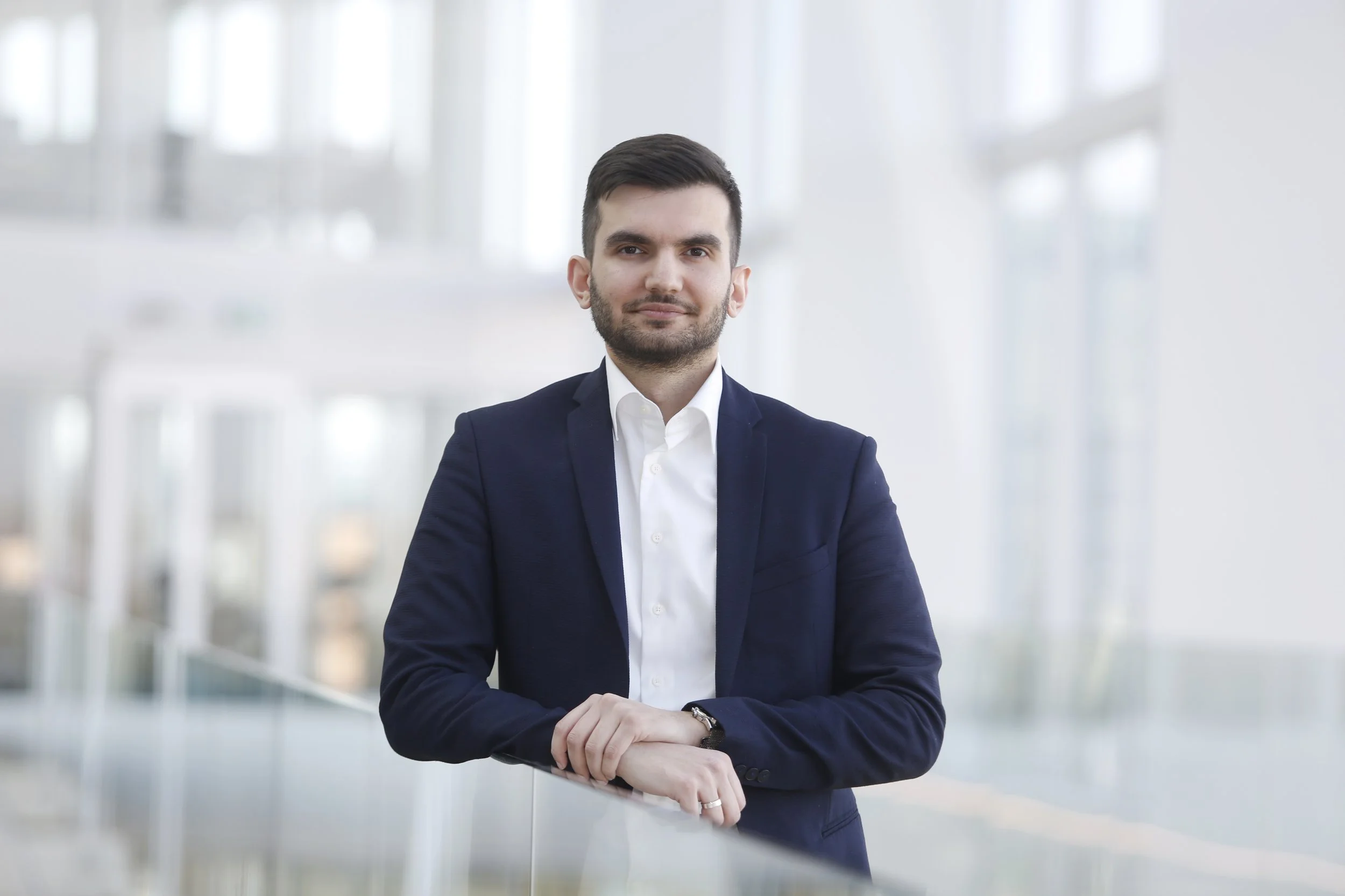 Zakelijke man in witte overhemd en donkerblauwe jas poseert in een modern kantoor met grote ramen.