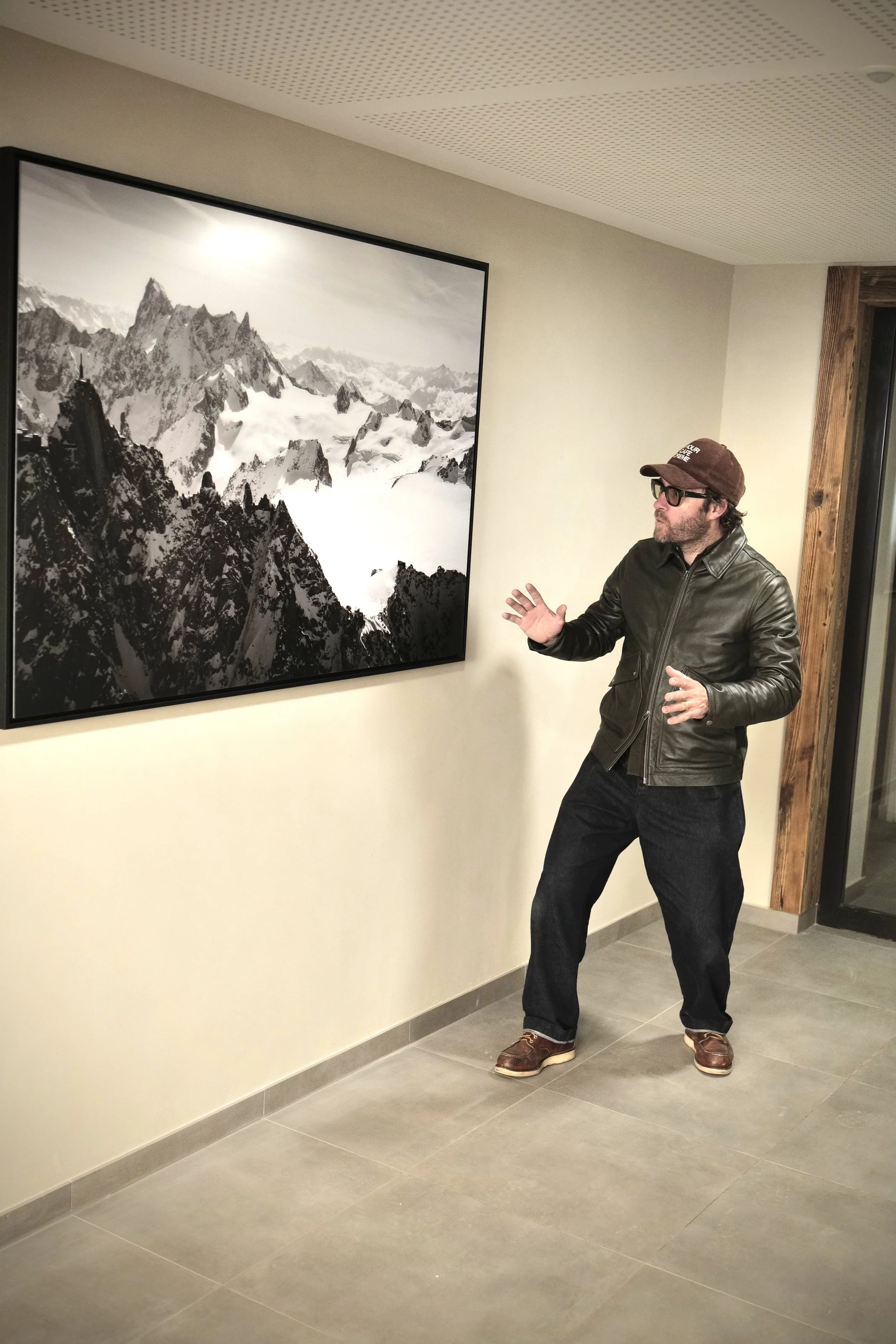 Un homme avec des lunettes et une casquette, portant une veste en cuir, se tient à côté d'une peinture ou d'une photographie en noir et blanc d'une chaîne de montagnes enneigées, dans une salle d'exposition ou une galerie.