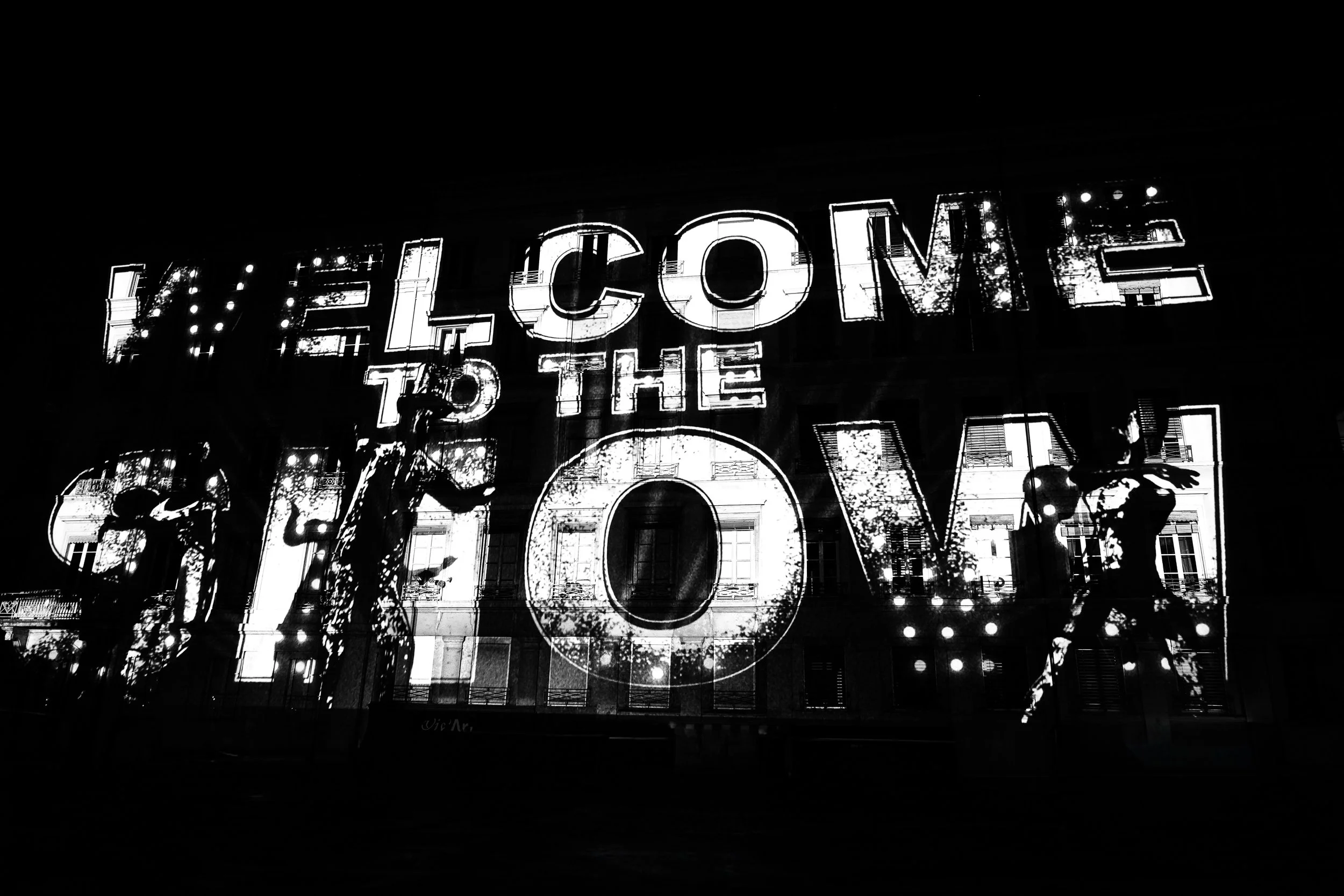 Un panneau lumineux en noir et blanc avec le texte « Welcome to the City » écrit en grandes lettres. La scène semble être prise la nuit, avec le reflet d'immeubles et d'arbres en arrière-plan.