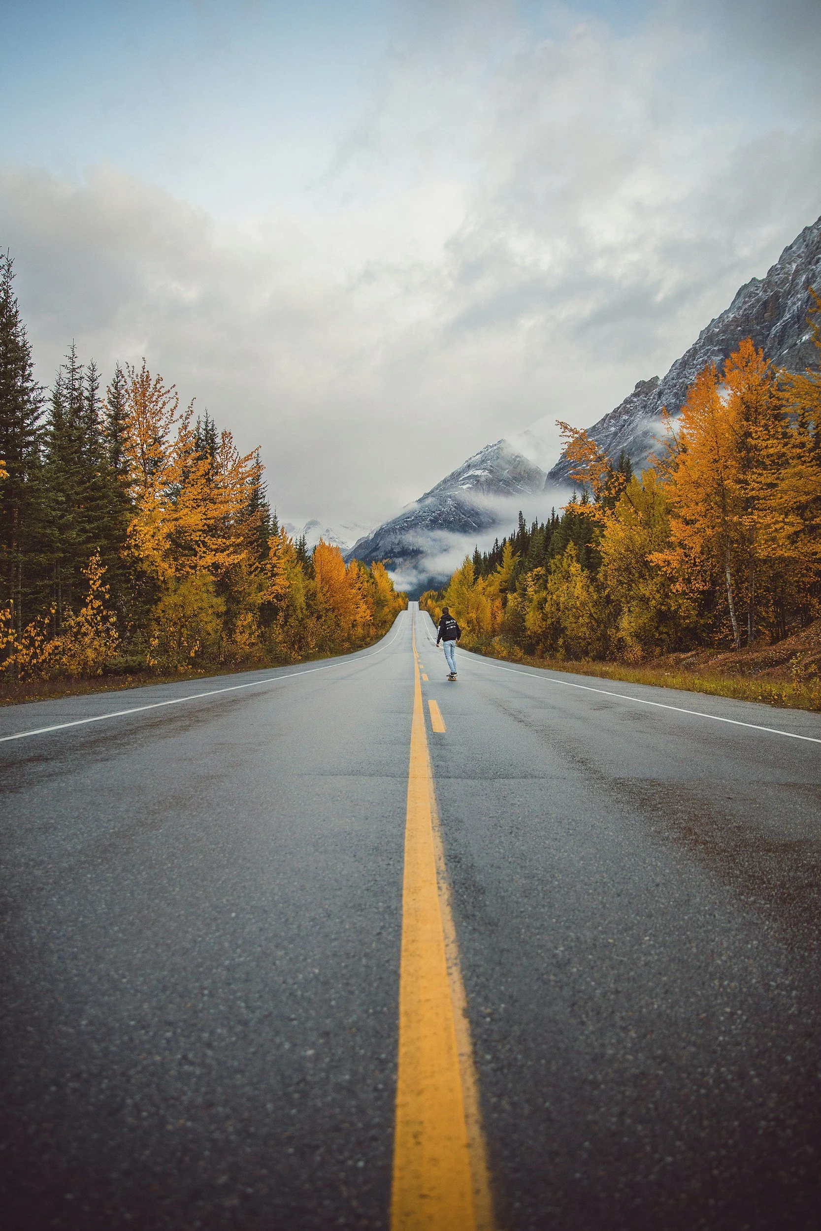 A person skateboarding on a long, straight mountain road lined with autumn trees, with mountains and clouds in the background.