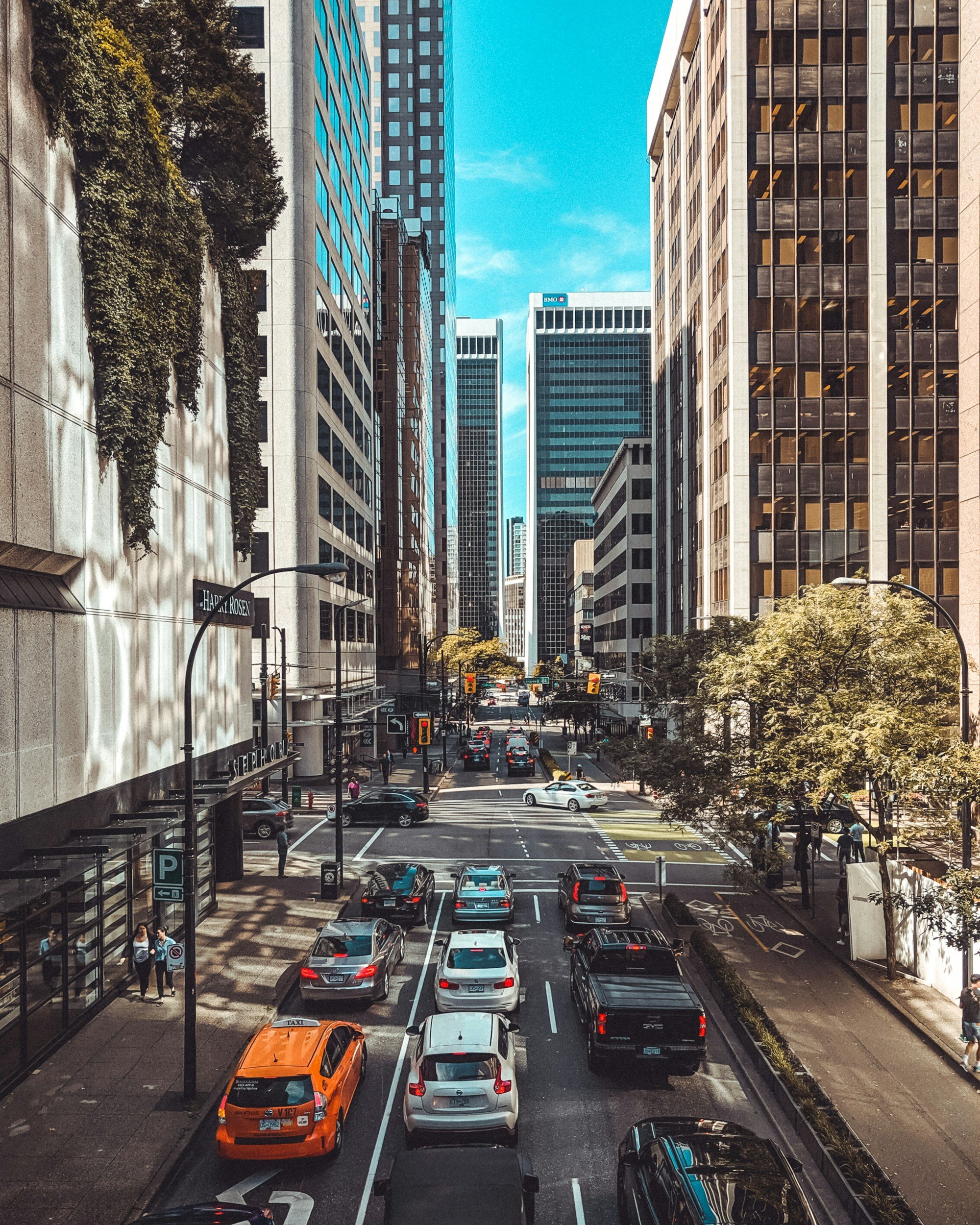 City street scene with tall office buildings, parked and moving cars, and pedestrians, under a bright blue sky.