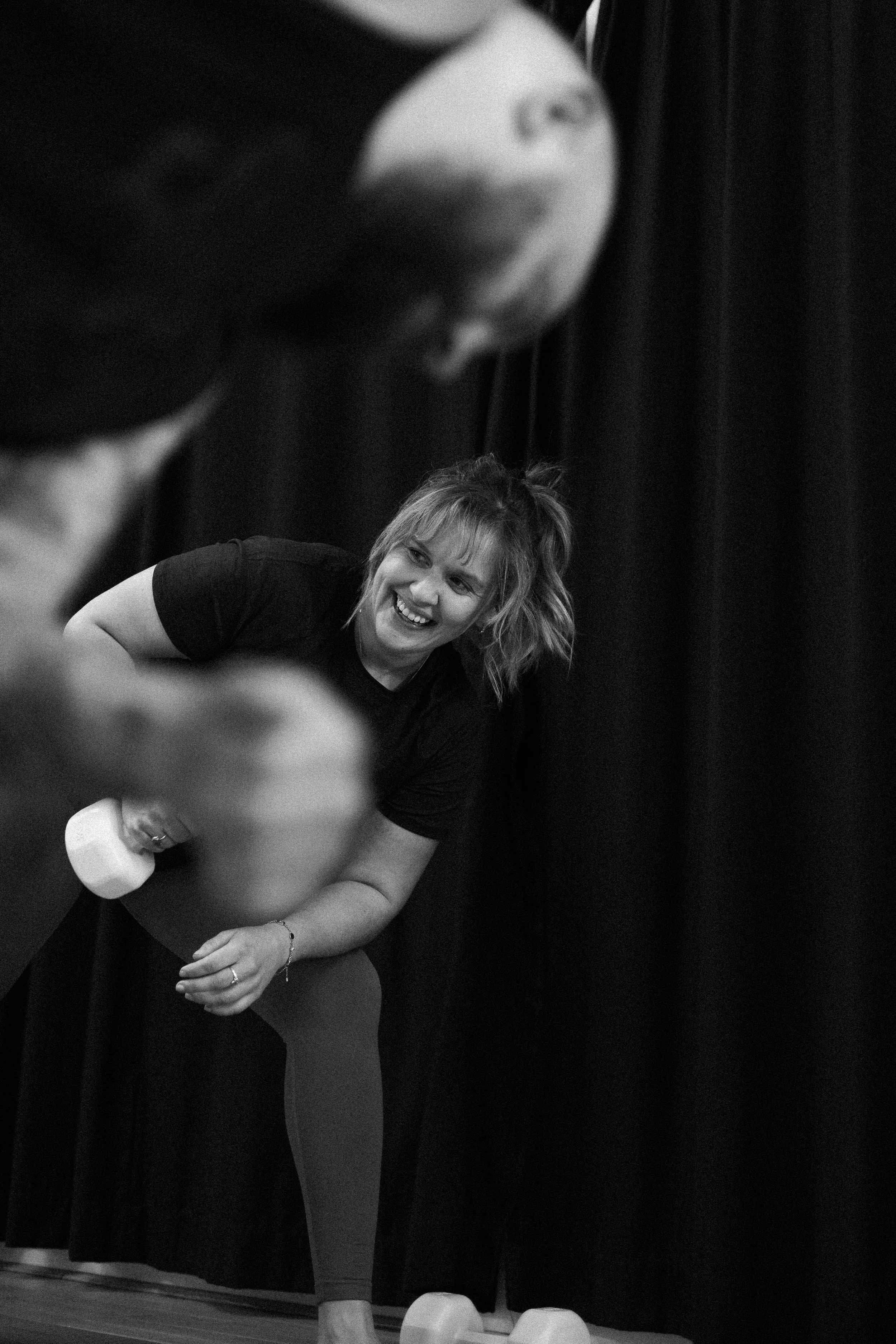 A woman with shoulder-length hair sitting on the floor smiling, with a young girl bouncing on a trampoline in front of her, dark curtain backdrop.