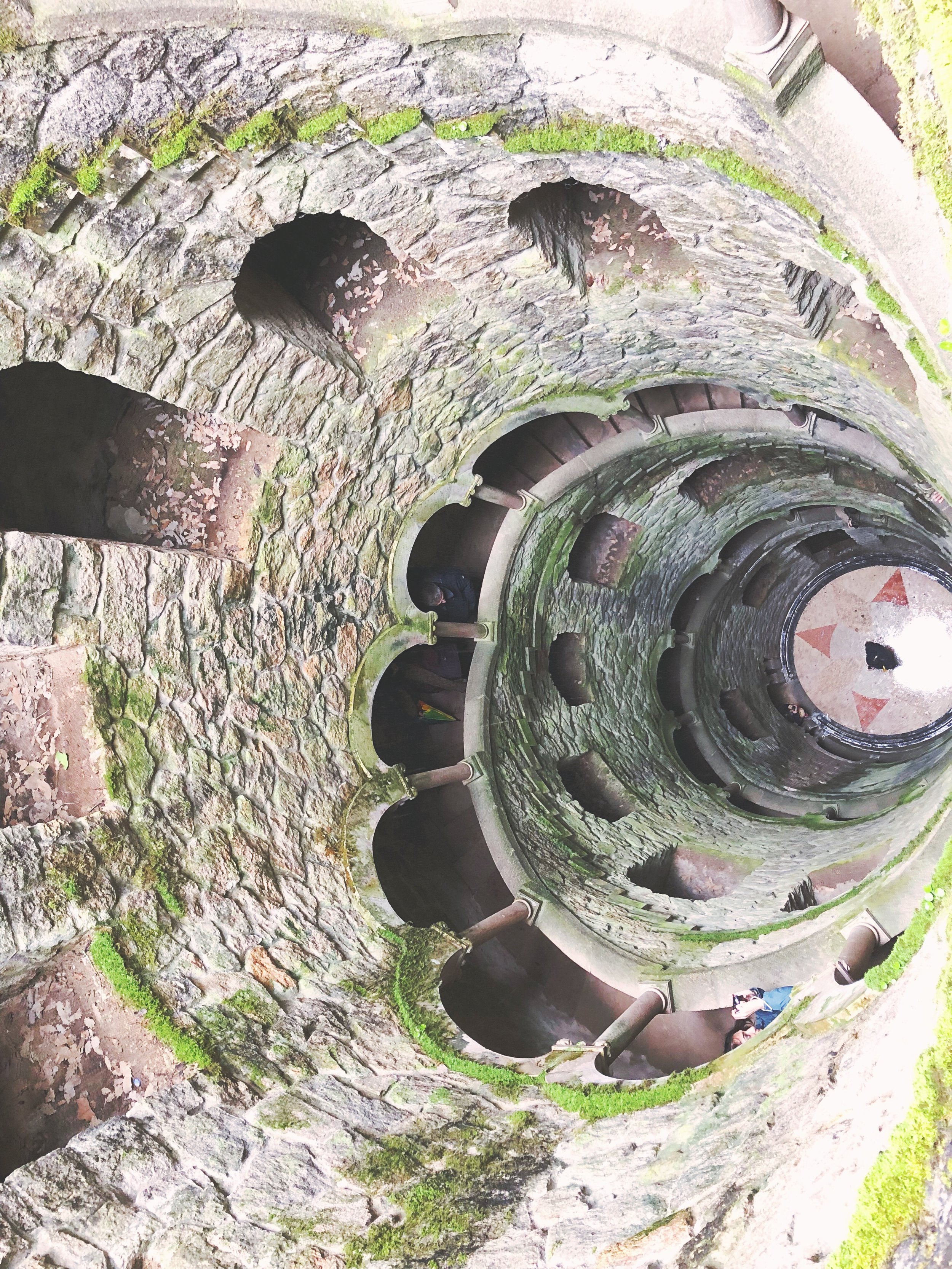 Spiral staircase made of moss-covered stone and concrete, with people walking on the stairs and holding onto the railing.