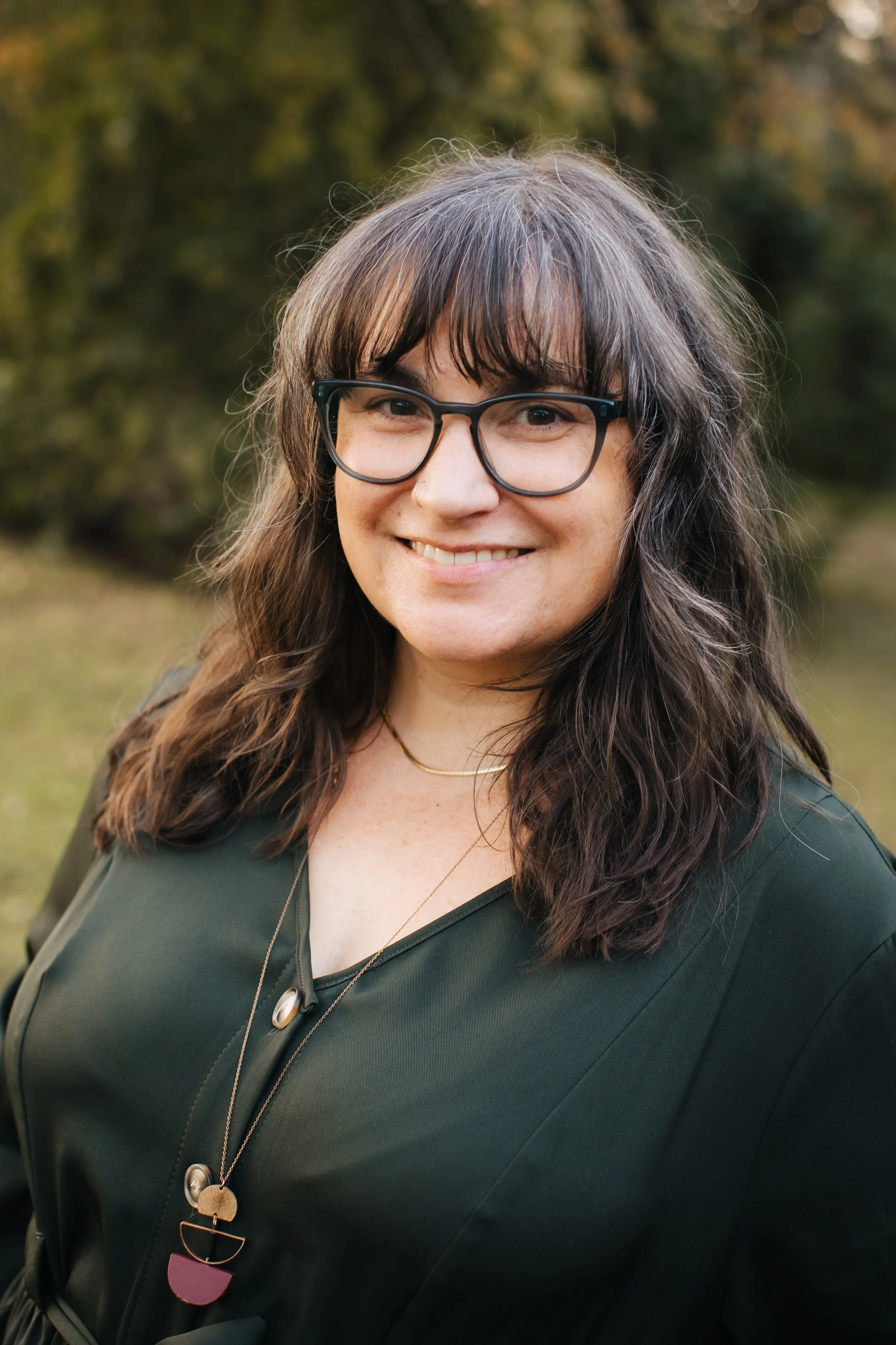 A woman with long dark hair and glasses smiling outdoors with a blurred green and brown background.