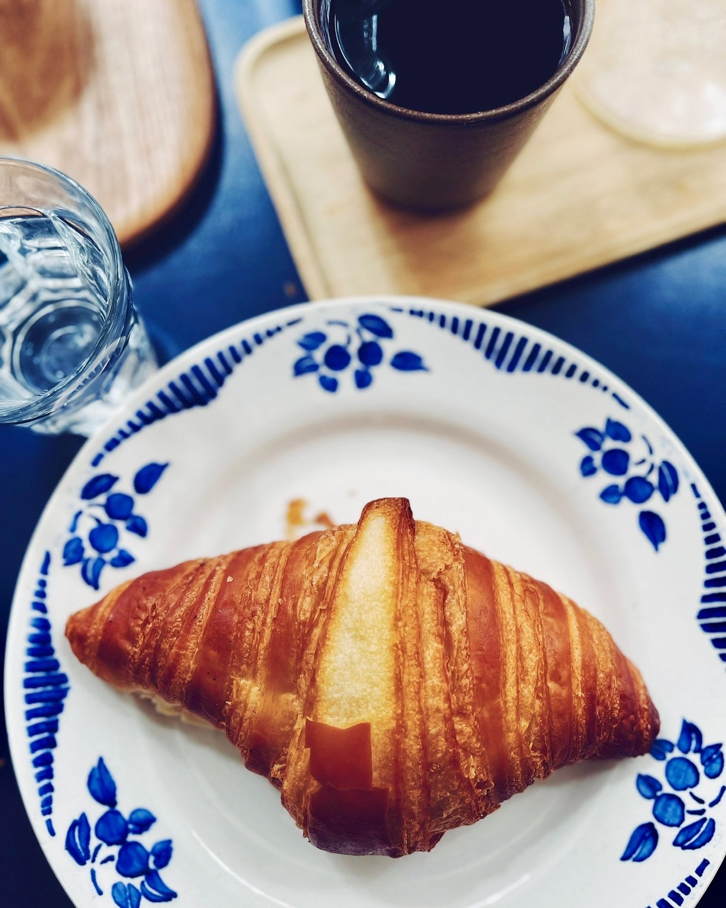 A croissant on a white plate with blue floral patterns, a glass of water, a black cup with a dark beverage, and a croissant on a wooden tray.