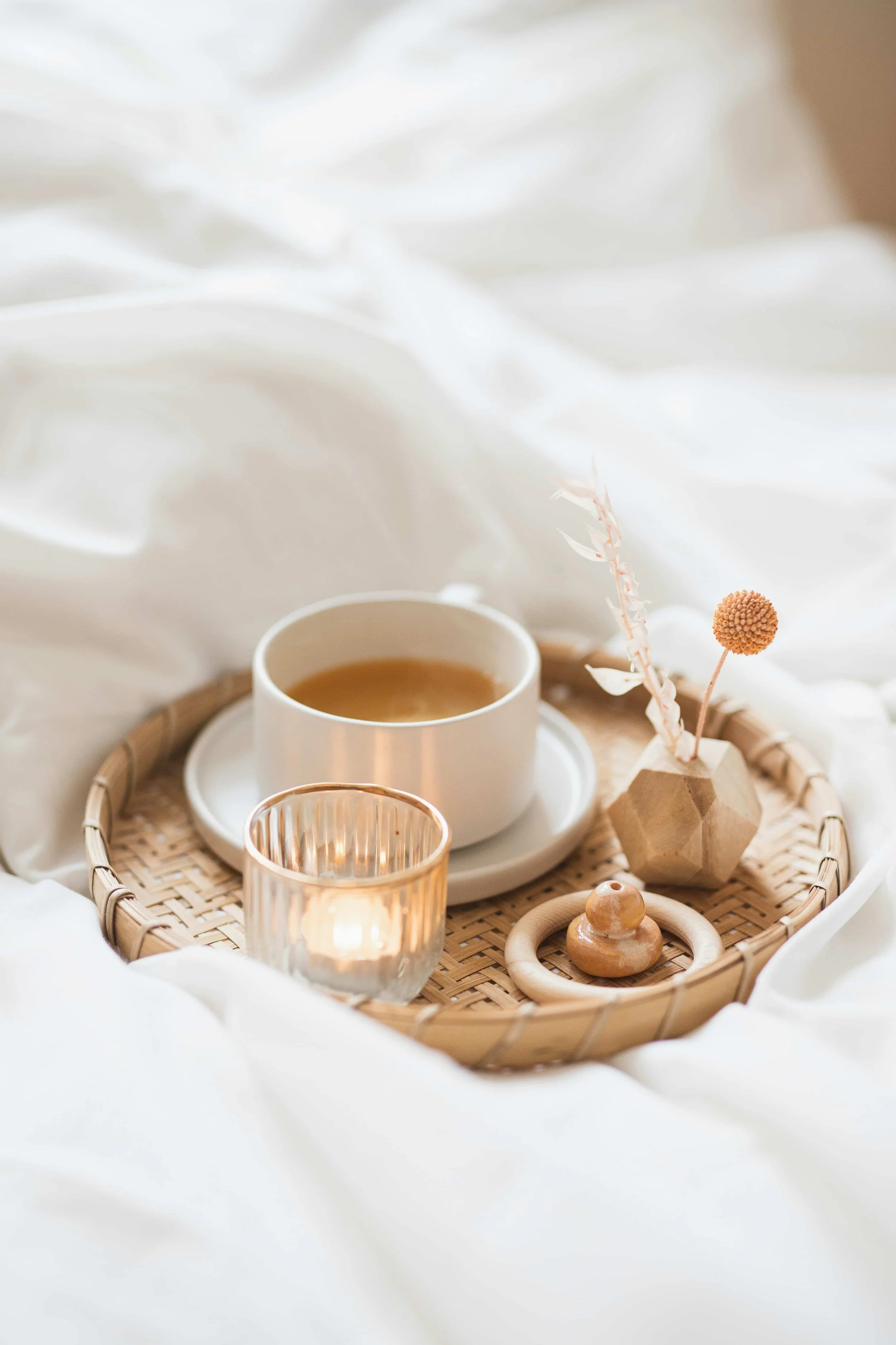 A tray with a cup of tea, a glass, a small candle holder, dried flowers, and wooden decorative items on a soft white fabric background.