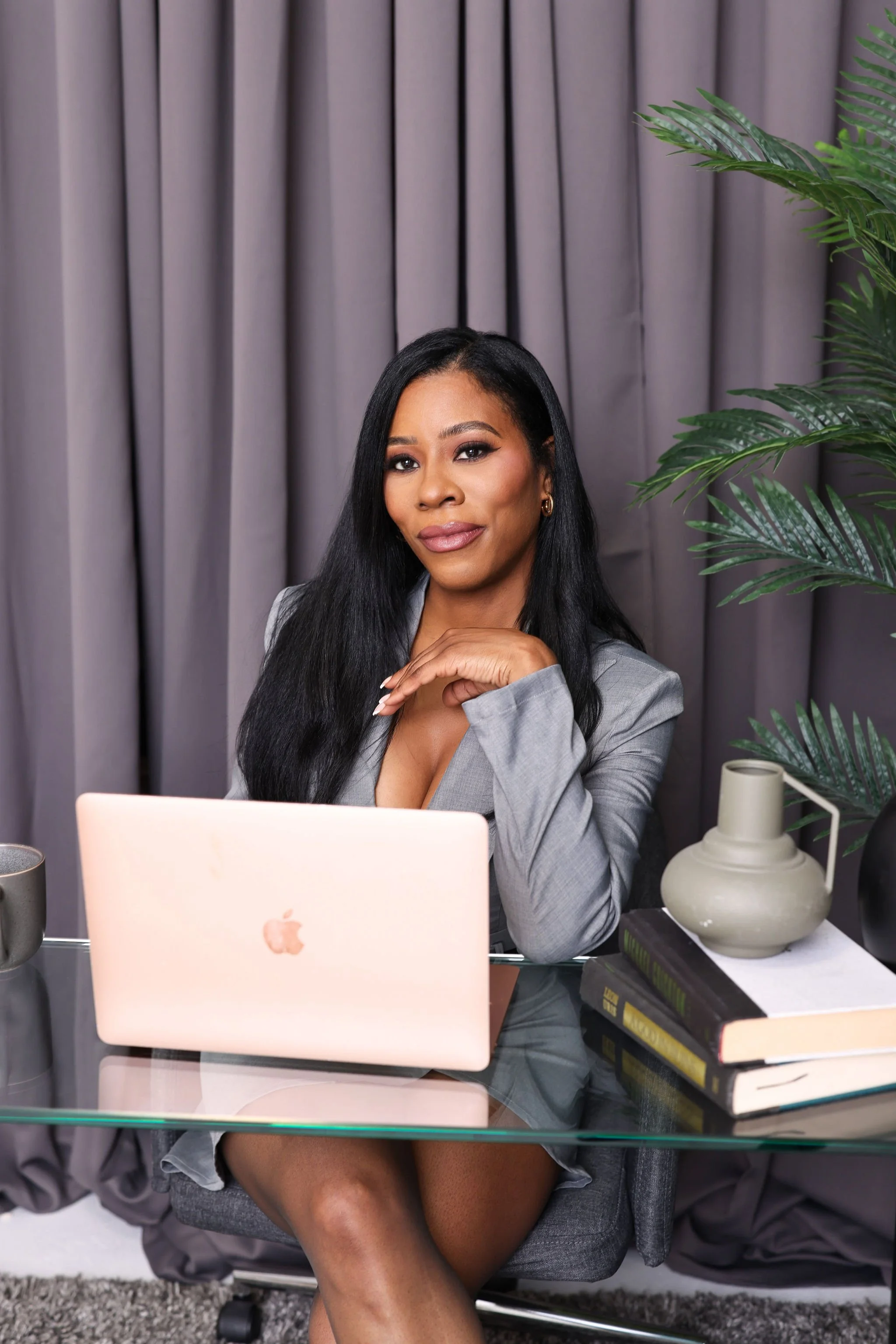A woman sitting at a desk with a laptop, books, and decorative items, including a plant and a lamp, in a professional setting with gray curtains in the background.