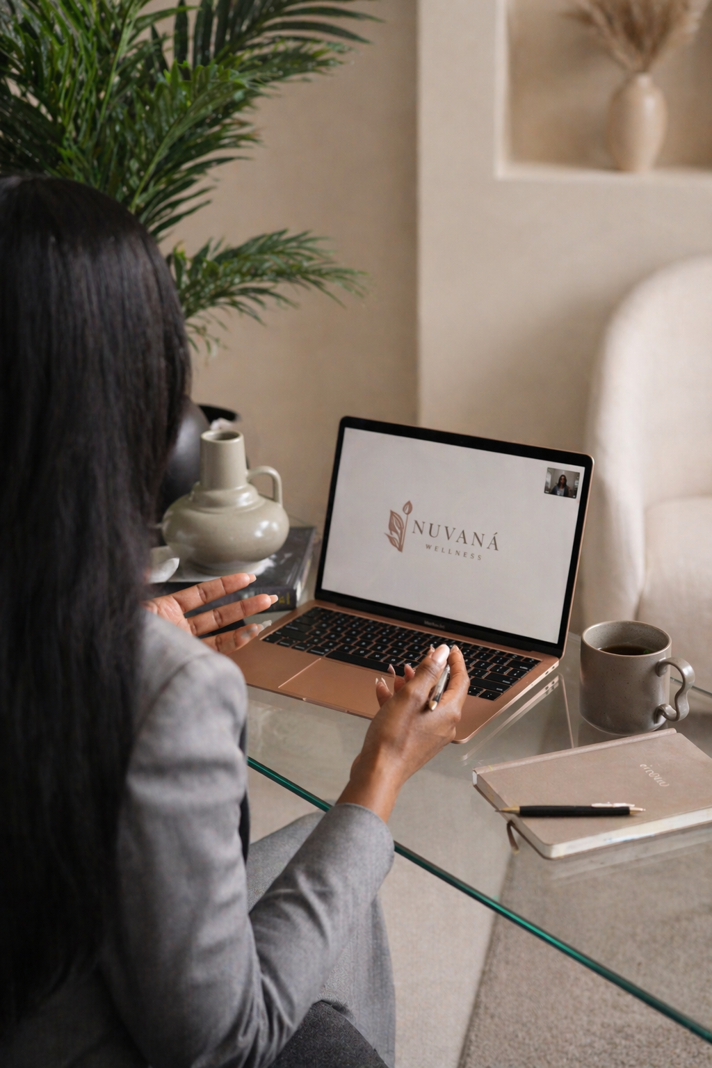 A woman sitting at a glass desk with a laptop that displays Nuvaná Wellness, a coffee mug, a closed notebook with a pen, a small book, and a decorative pitcher. There is a large green plant behind her and a beige wall with a built-in niche containing a vase with dried branches.