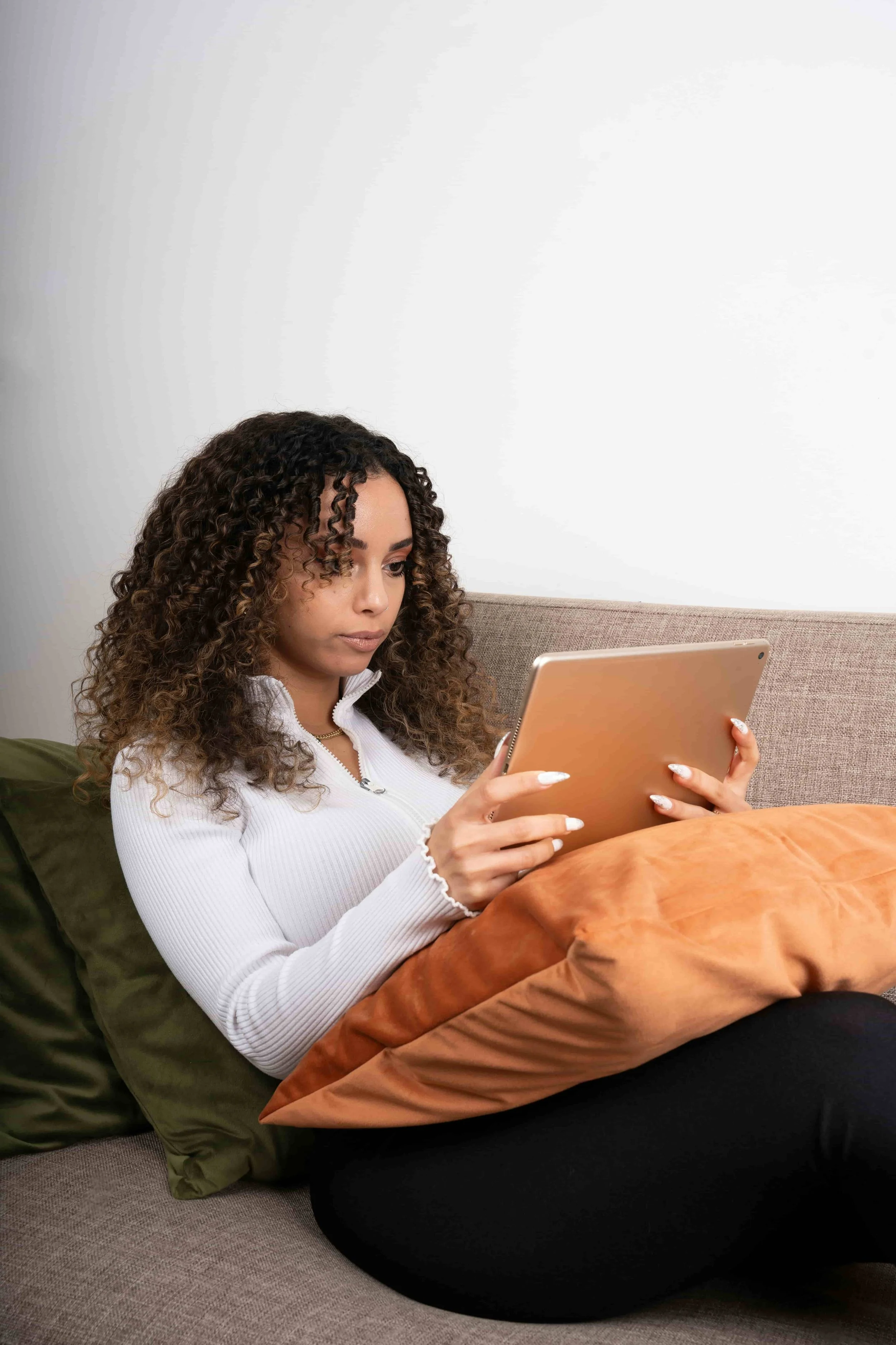 A young woman with curly hair sitting on a couch, looking at a tablet for a telehealth session, with brown and green cushions behind her and a white wall in the background.