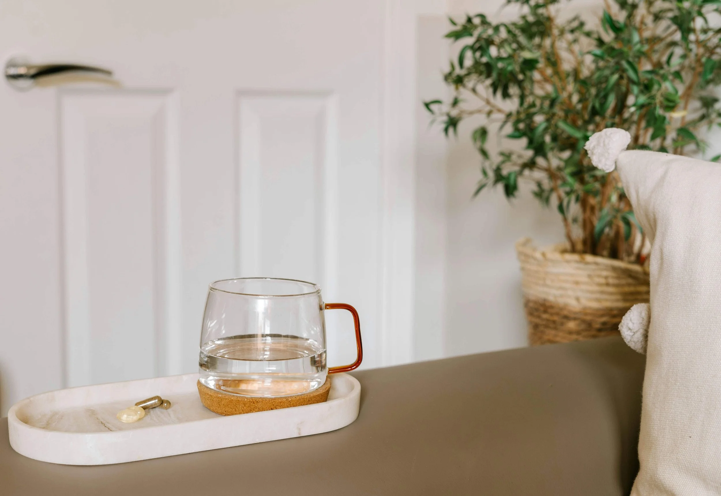 A clear glass mug filled with water, placed on a cork coaster on a white marble tray, with jewelry beside it, set on a surface with a potted plant and a white cloth in the background.