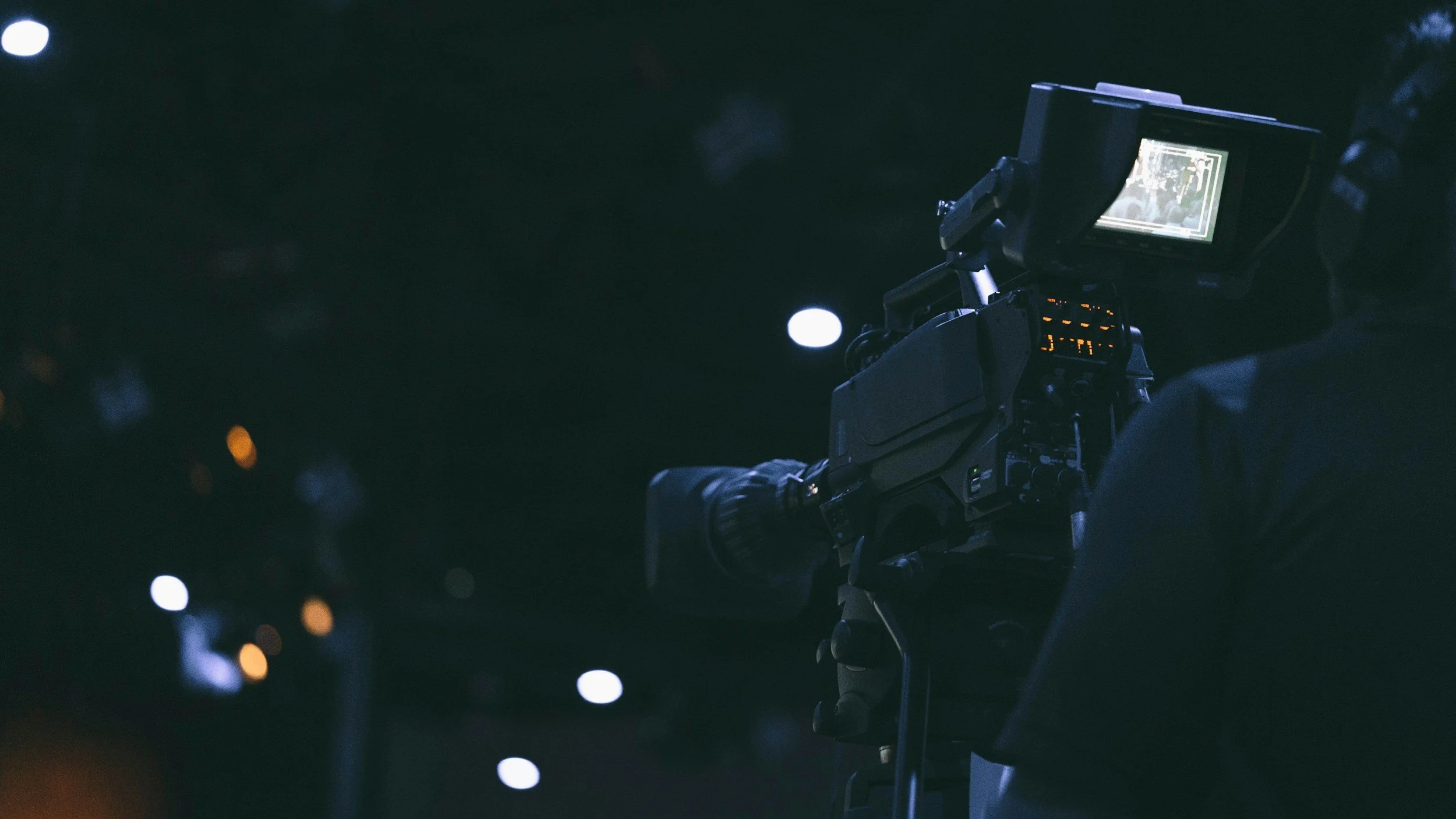 A camera operator filming at night, with city lights blurred in the background.