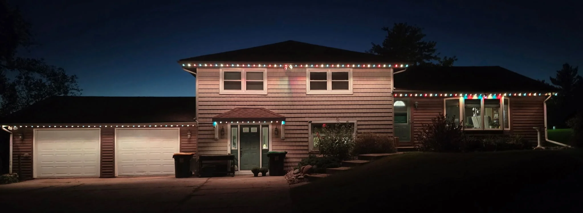 Nighttime view of a two-story house decorated with colorful permanent lighting along the roofline, with a garage and a front door, surrounded by trees and a dark sky.