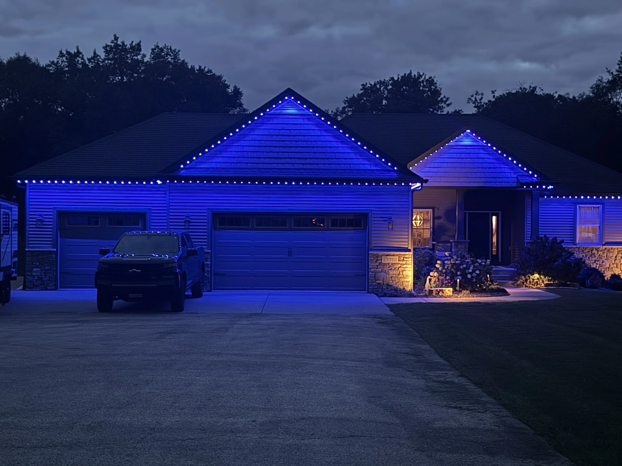 A house at dusk decorated with blue LED permanent lights outlining the rooflines and eaves. The house has a garage, a window with a warm light, and a well-maintained lawn with flowers in front.