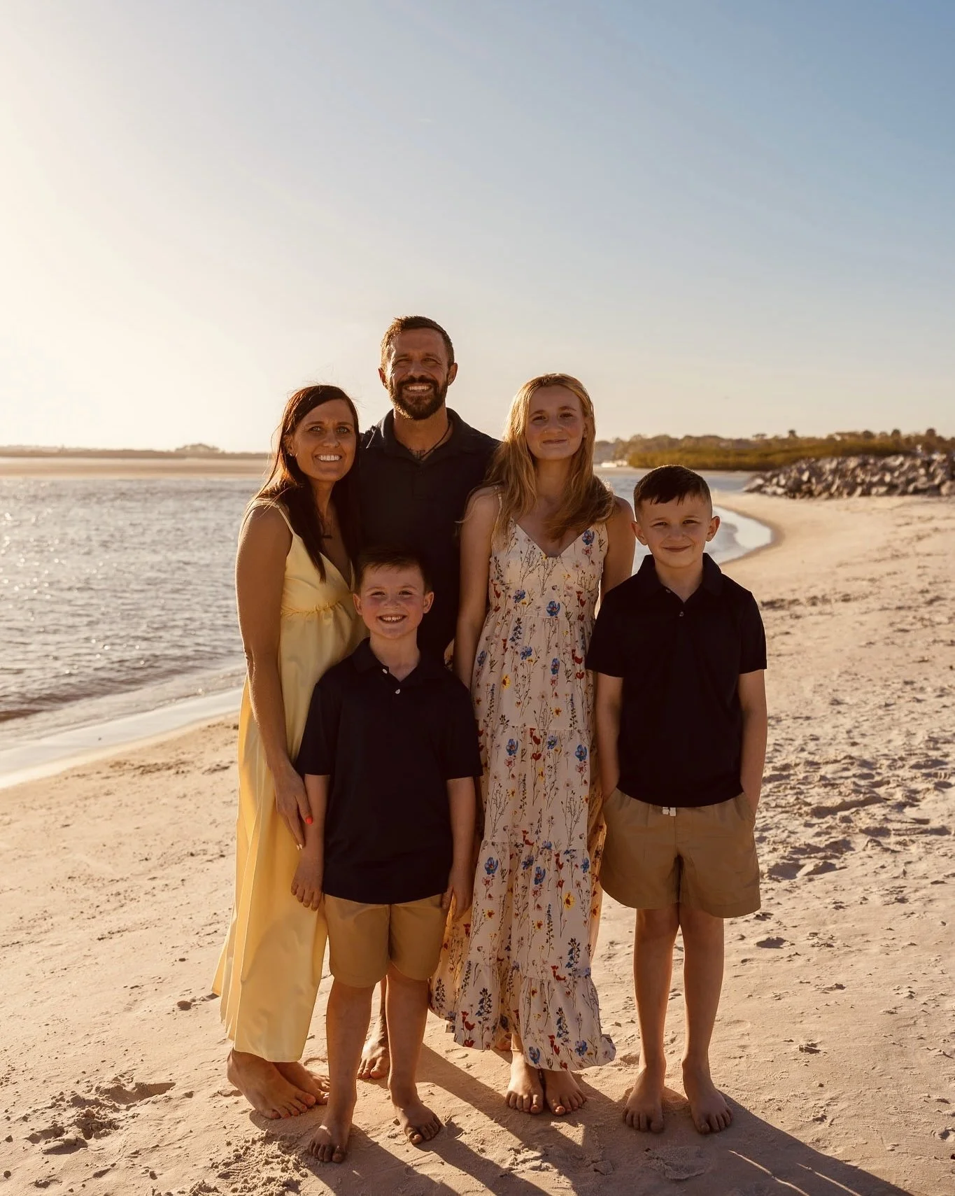 A family of five standing on a beach at sunset, with a calm body of water and a rocky shoreline in the background. The family members are smiling and dressed in casual summer clothing.