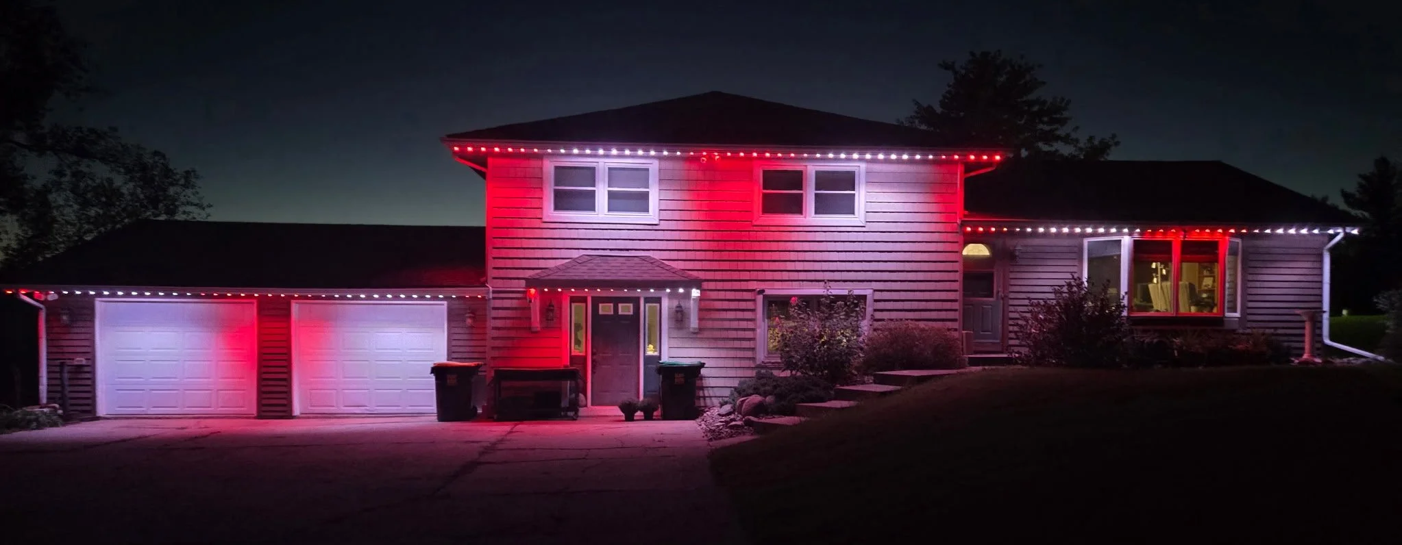 Two-story house decorated with red and white permanent lighting at night, with a two-car garage, shrubs, and trees in the background.