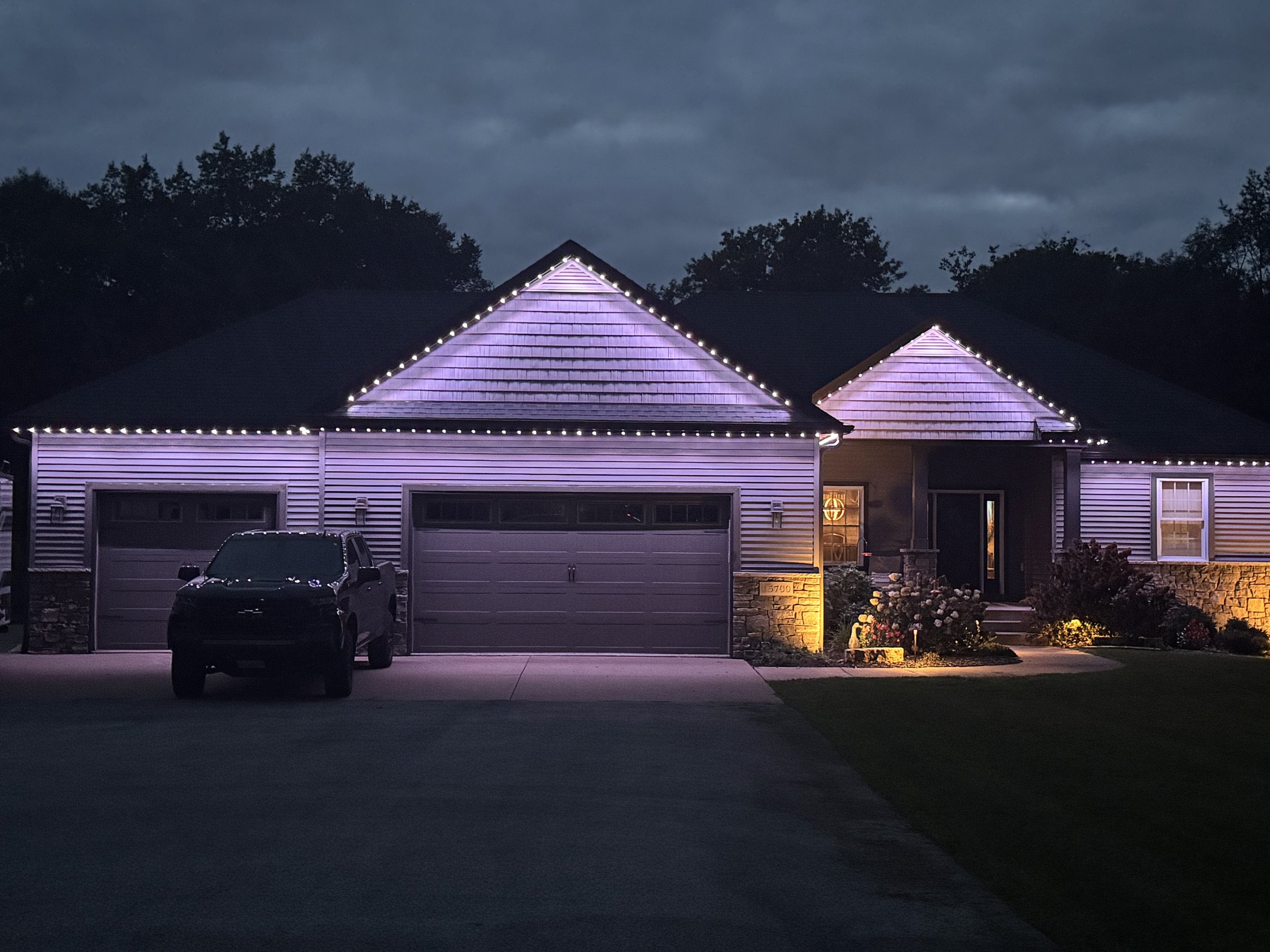 A house decorated with permanent lighting on the roof at night. The house has a two-car garage, a front porch with a small garden, and a dark sky with clouds overhead.