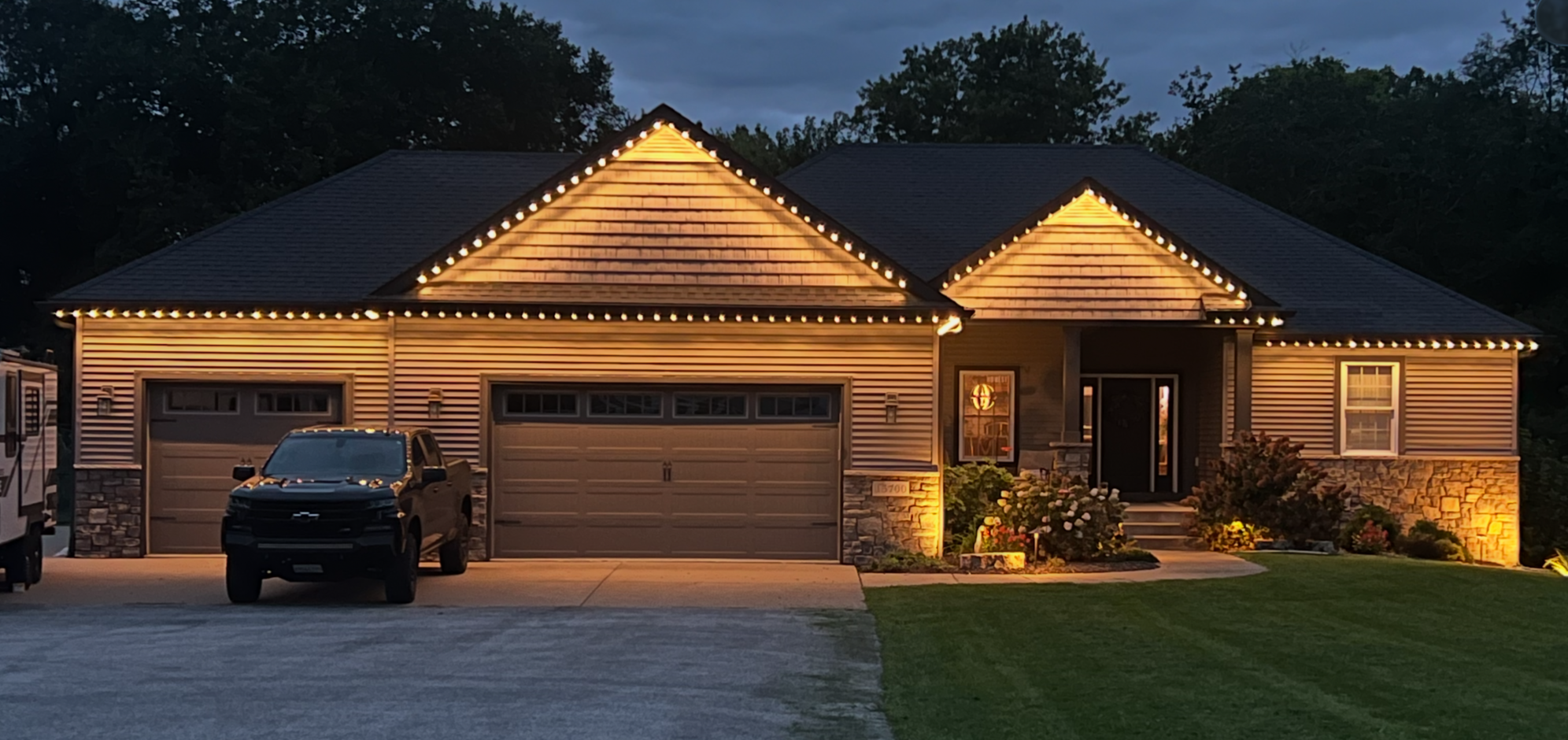 A house decorated with permanent lighting along the roofline, during dusk, with a driveway in front and a black truck parked outside. The house features a garage, a front door with a window, and landscaped garden with flowers and bushes.