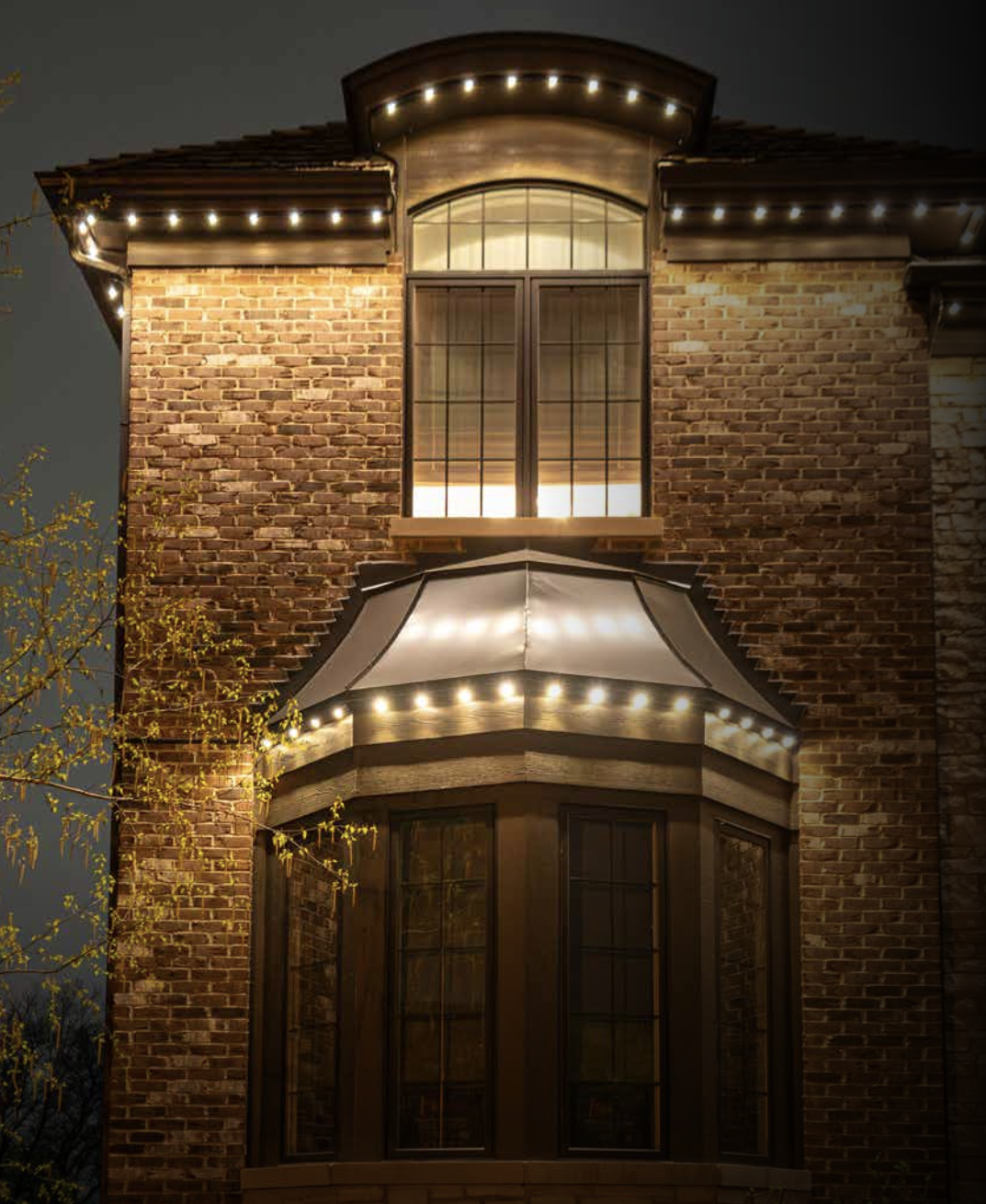 Illuminated brick house at night with string lights along roofline and bay window