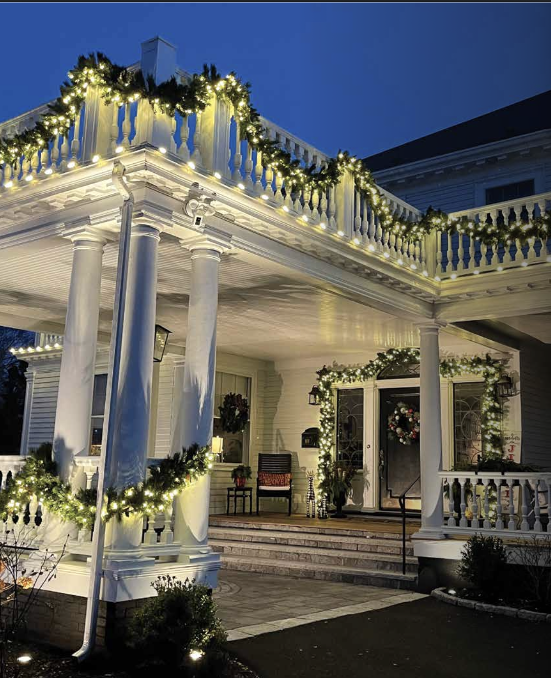 A house decorated with Christmas greenery and string lights on the porch and upper balcony during the evening.