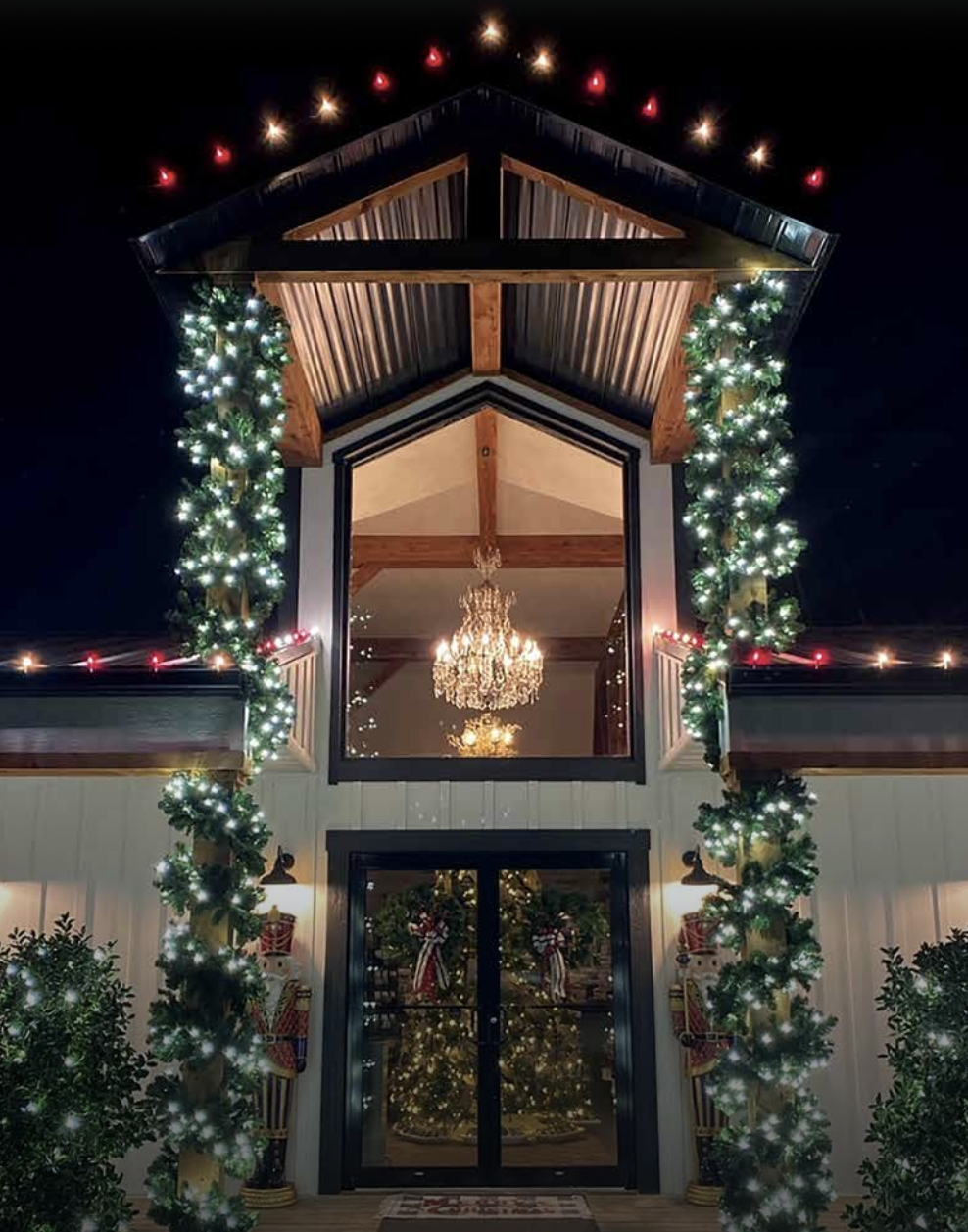 Festively decorated barn with green garlands and white lights framing the entrance, a Christmas tree with lights visible through glass doors, and chandeliers hanging inside, topped with Christmas Nutcracker figures on either side.