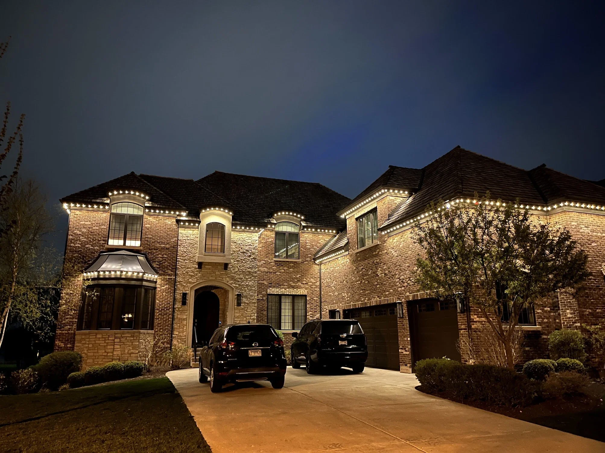 Nighttime view of a large brick house with string lights outlining the roof and windows, and a driveway with parked cars in front.